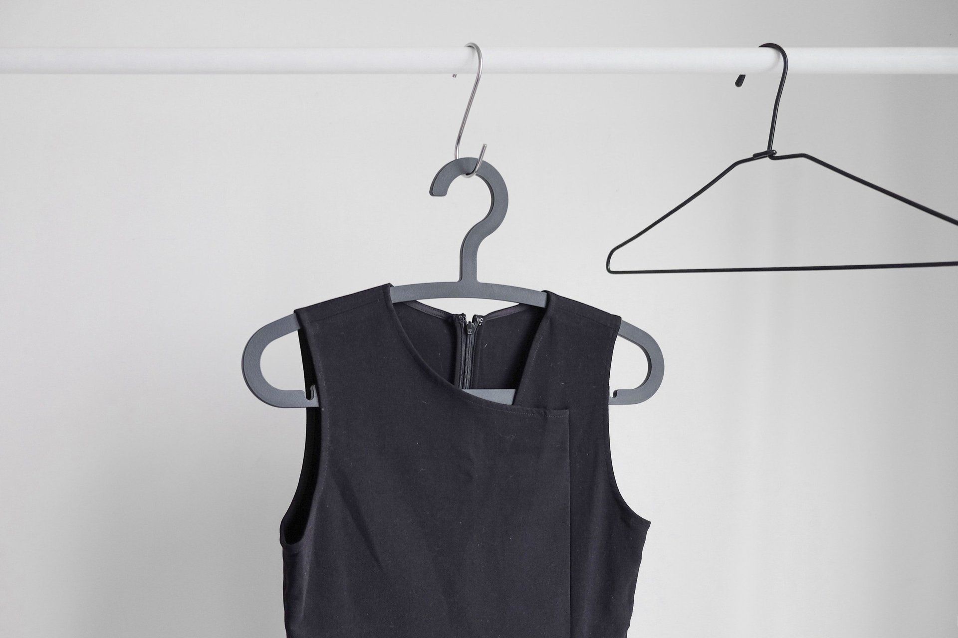 Een zwart mouwloos topje hangt aan een hanger, met daarnaast nog een lege hanger aan een witte stang tegen een lichte muur.
