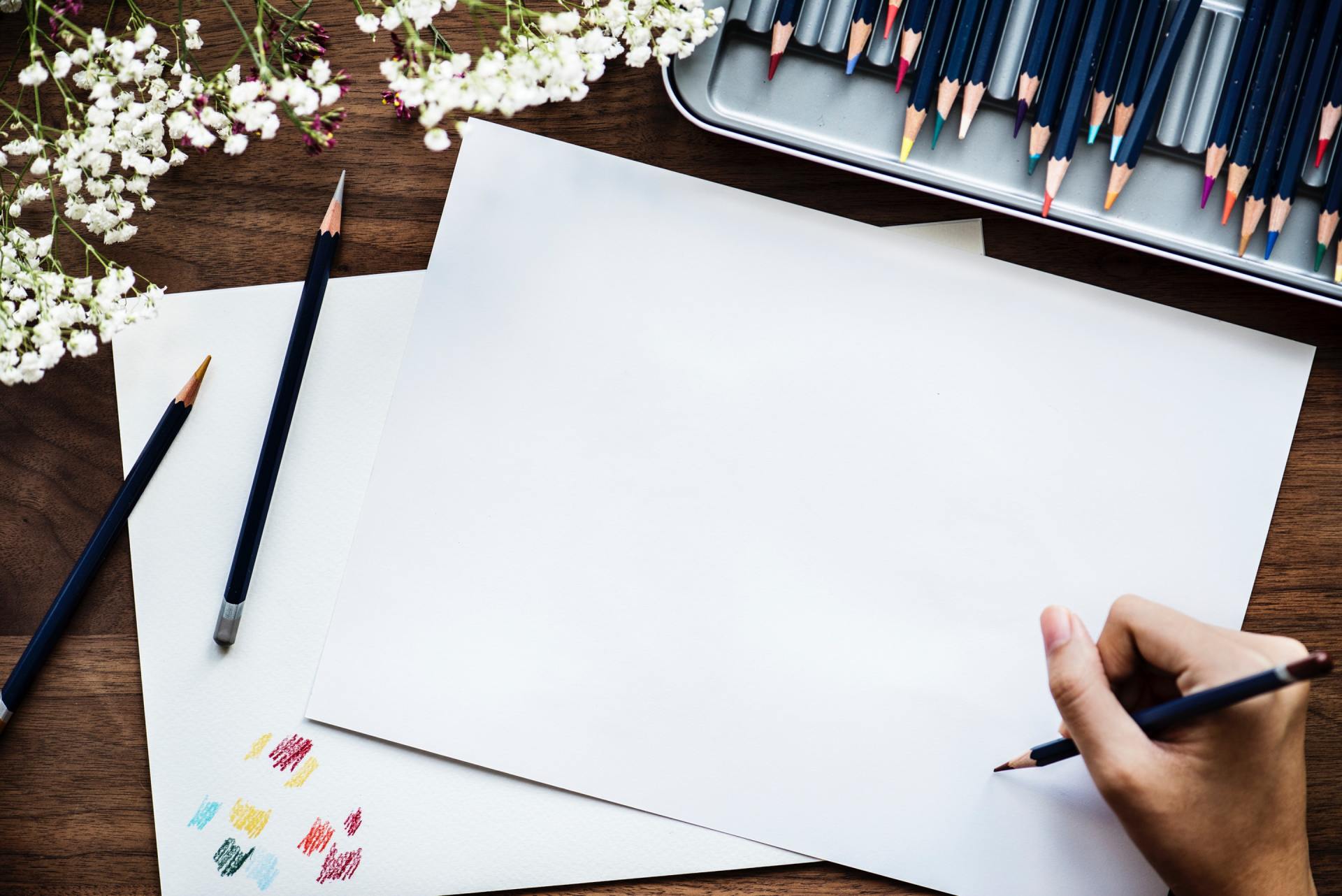 Hands sketching on blank paper with pencils and paint swatches on a wooden desk