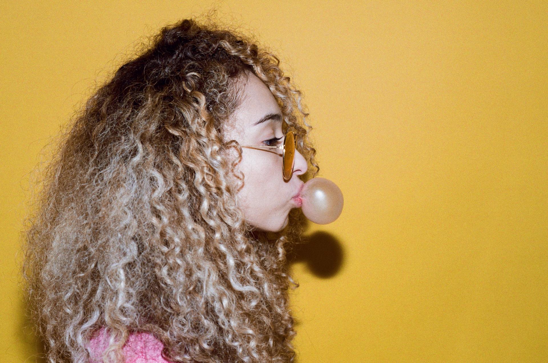 A woman with curly hair is blowing a bubble of gum.