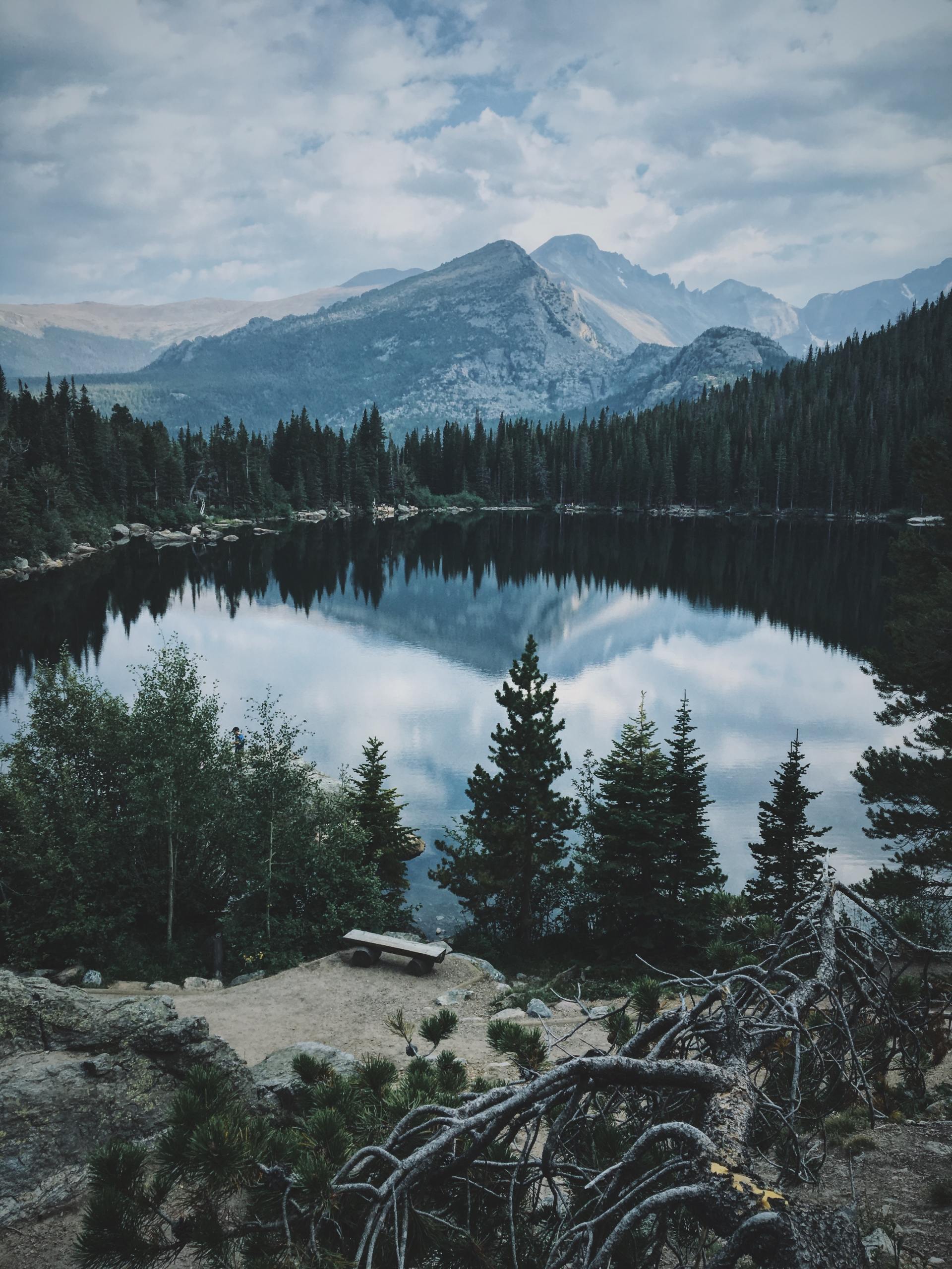 Calm lake reflects mountains and sky, surrounded by dark green trees under cloudy skies.