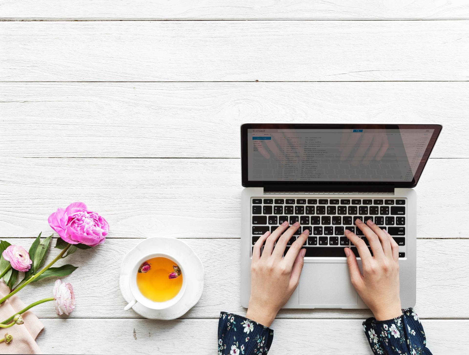 Person typing on laptop with tea and pink flowers on a white wood table.
