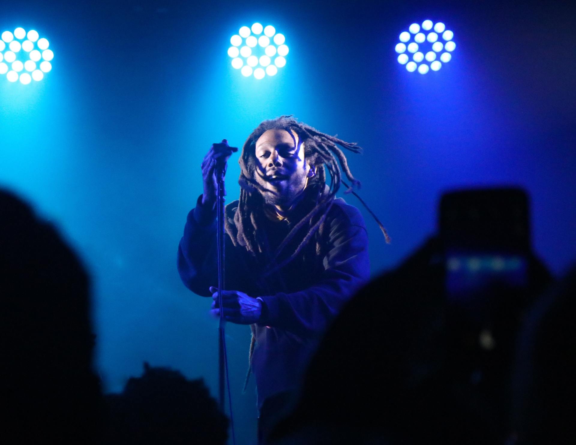 A person with dreadlocks performing on a stage under blue spotlights, holding a microphone, with a dark, blurred crowd.