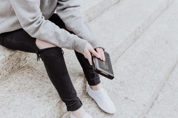 Een persoon in een grijze sweater en een zwarte gescheurde spijkerbroek, met een boek in de hand, zit op een stenen trap.