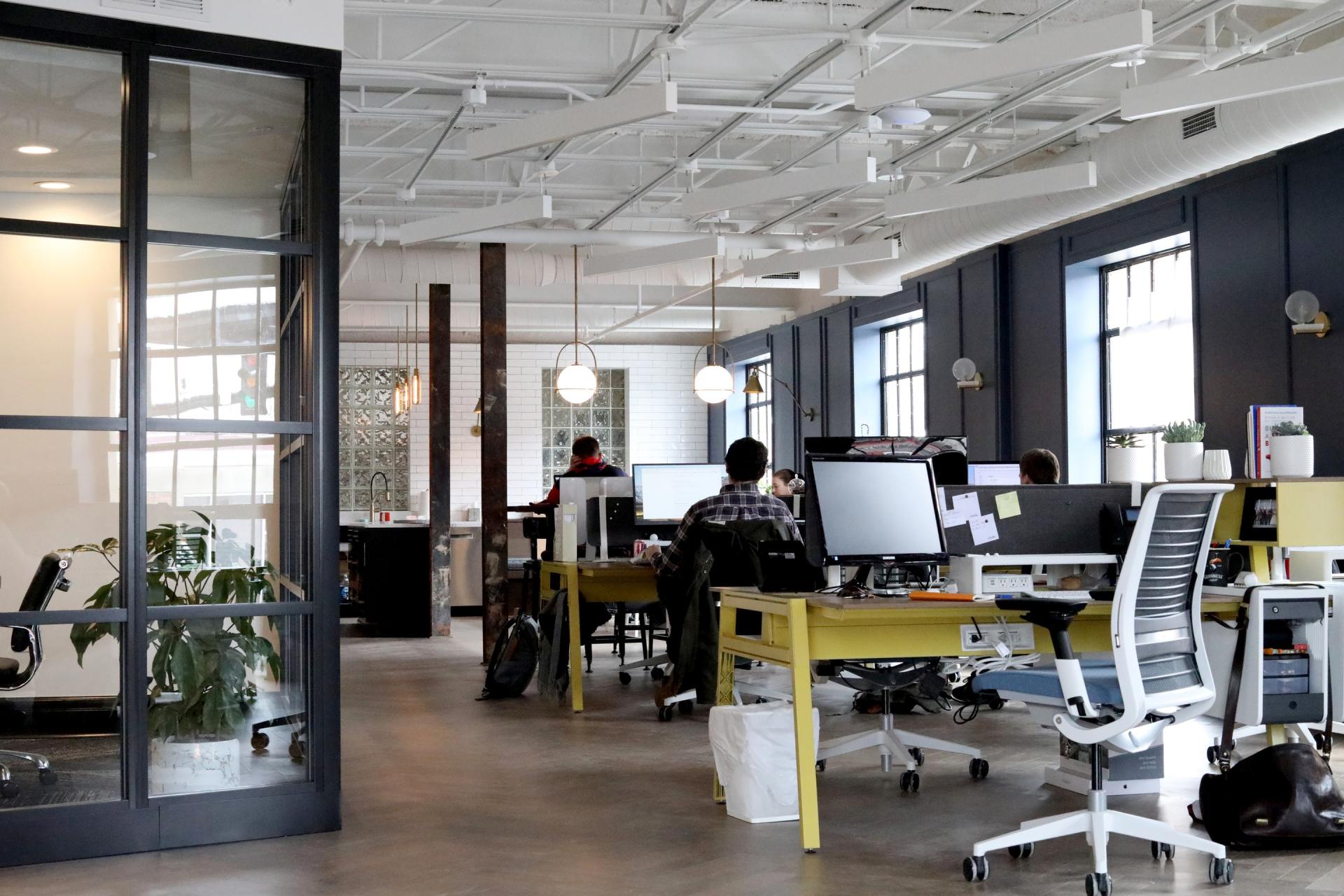 Modern office with people working at desks. Open space with exposed ceiling, large windows, and glass-paneled door.
