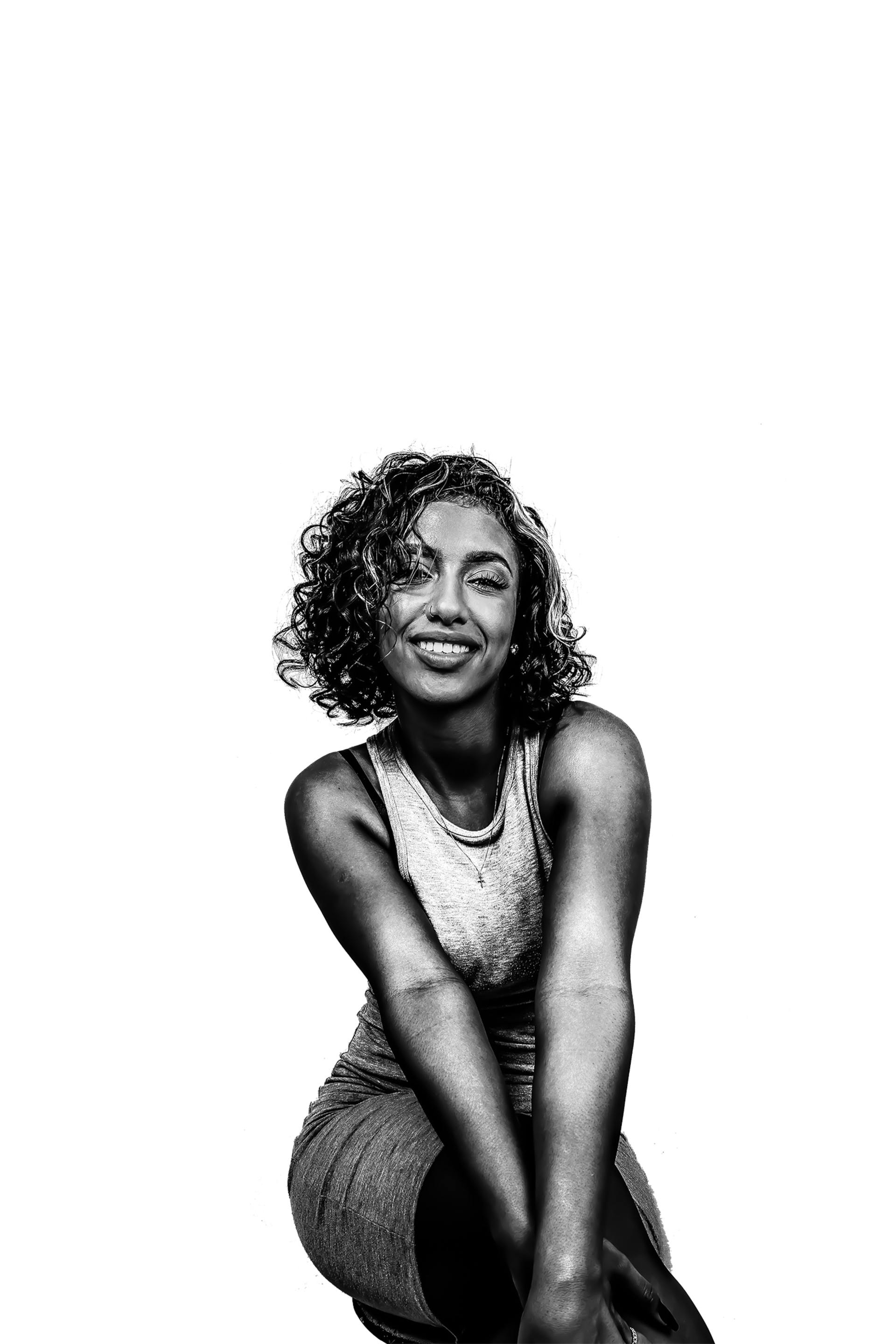 Black-and-white portrait of a smiling person with curly hair, sitting cross-legged against a white background