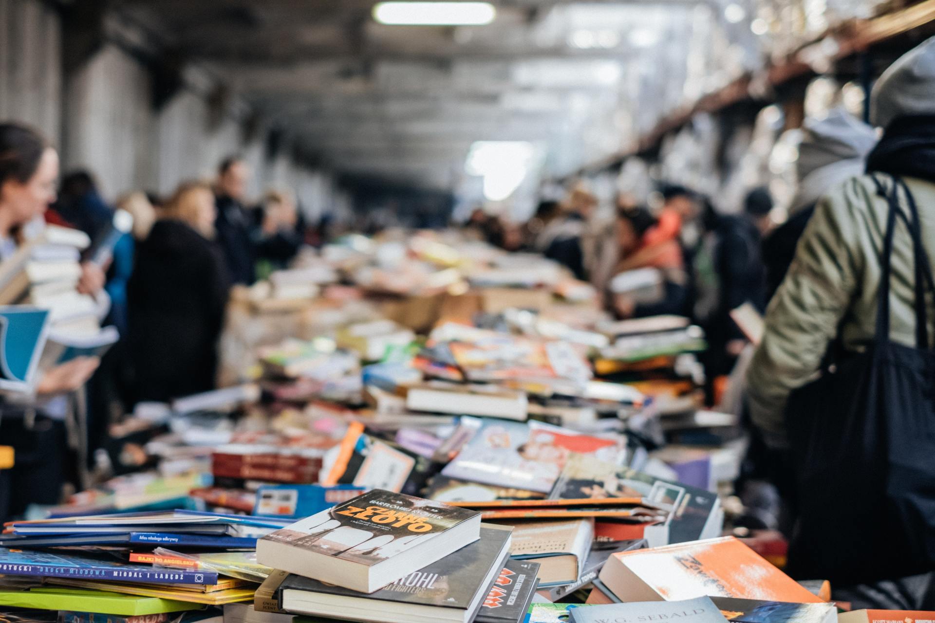 Books piled on tables at an outdoor book market; people browse, selecting books.