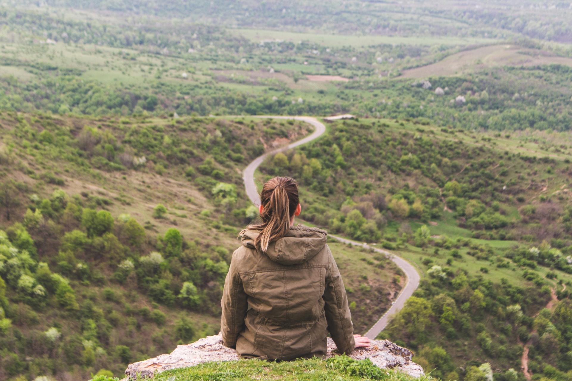 A woman is sitting on top of a hill looking at a road.