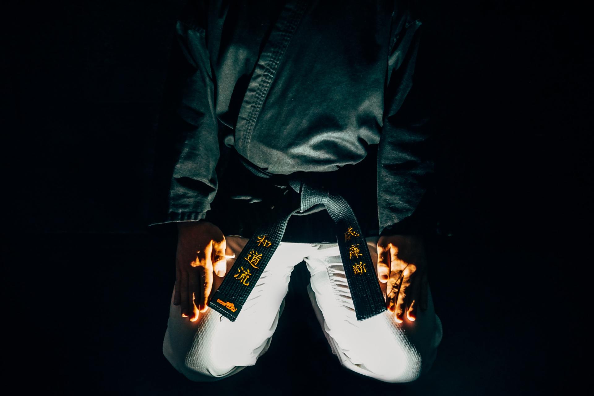 Person kneeling in a black martial arts uniform, hands glowing with orange light, black belt with gold characters.
