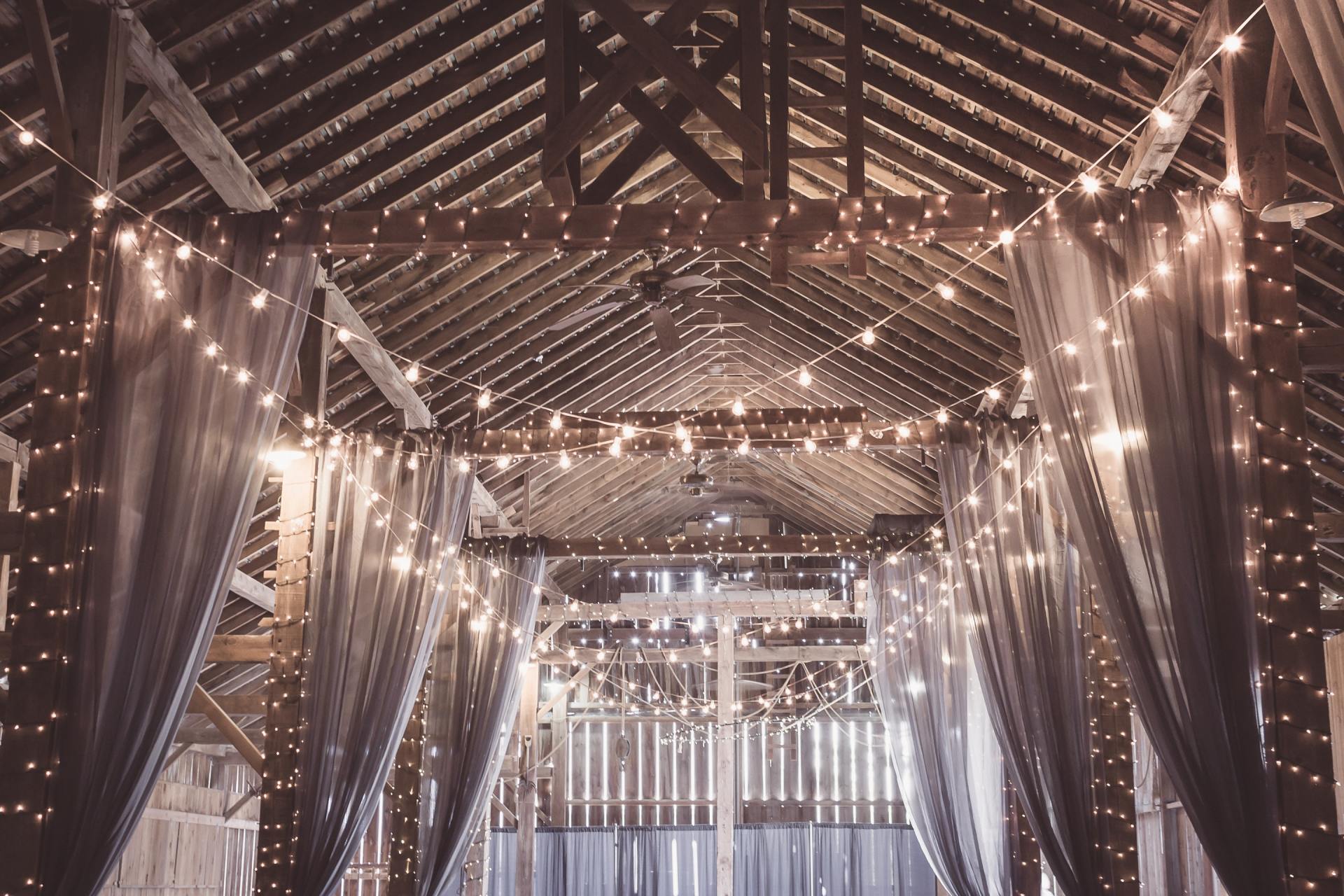 Rustic barn interior decorated with string lights and sheer drapes for a warm, inviting atmosphere.