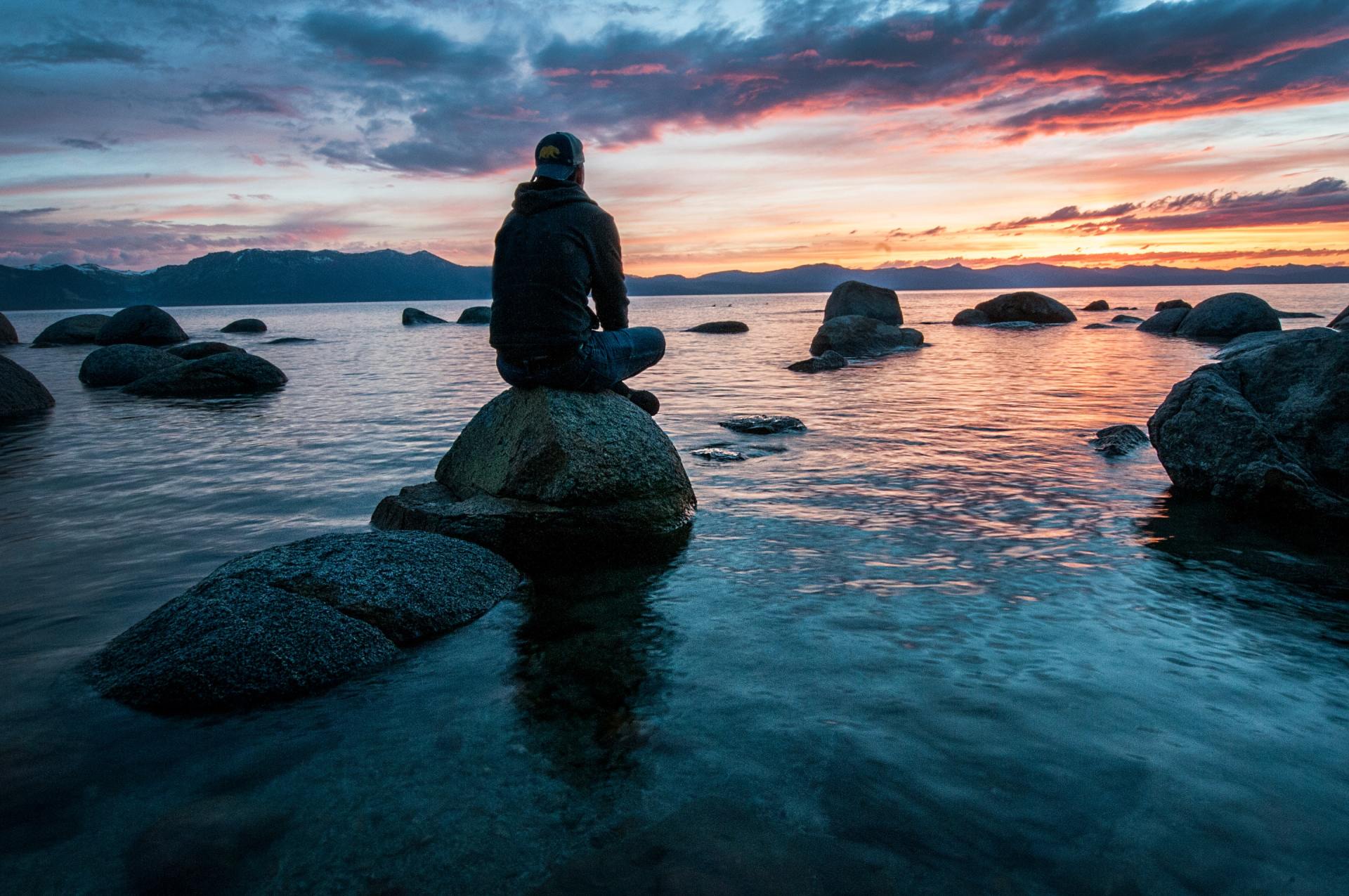 Person sitting on a rock in a calm sea at sunset, with scattered rocks and a colorful sky