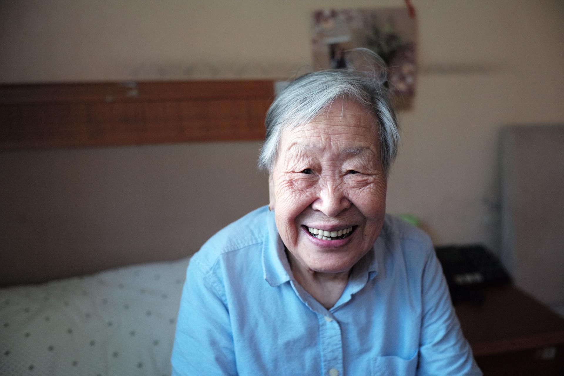 An elderly woman is smiling while sitting on a bed.