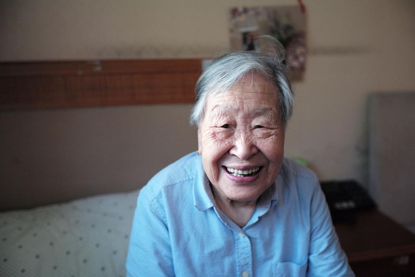 An elderly woman is smiling while sitting on a bed.