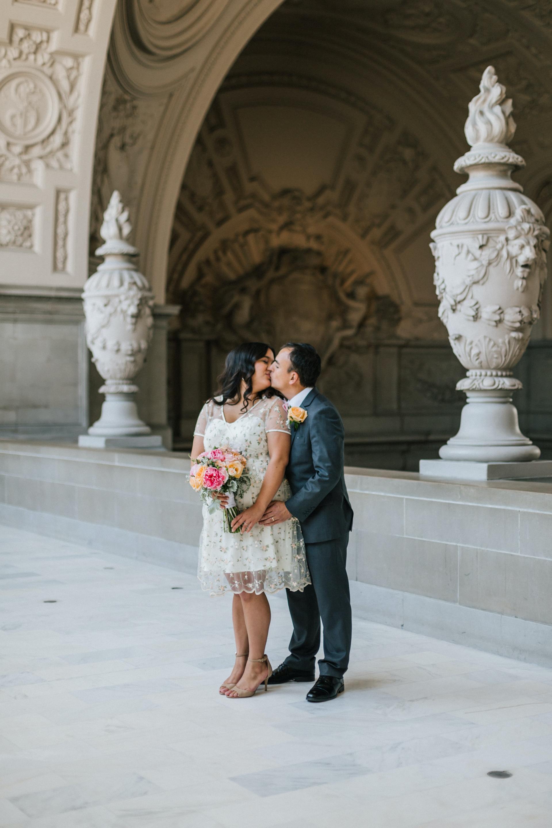 Couple kissing during a wedding ceremony in an ornate building. The bride holds a colorful bouquet.