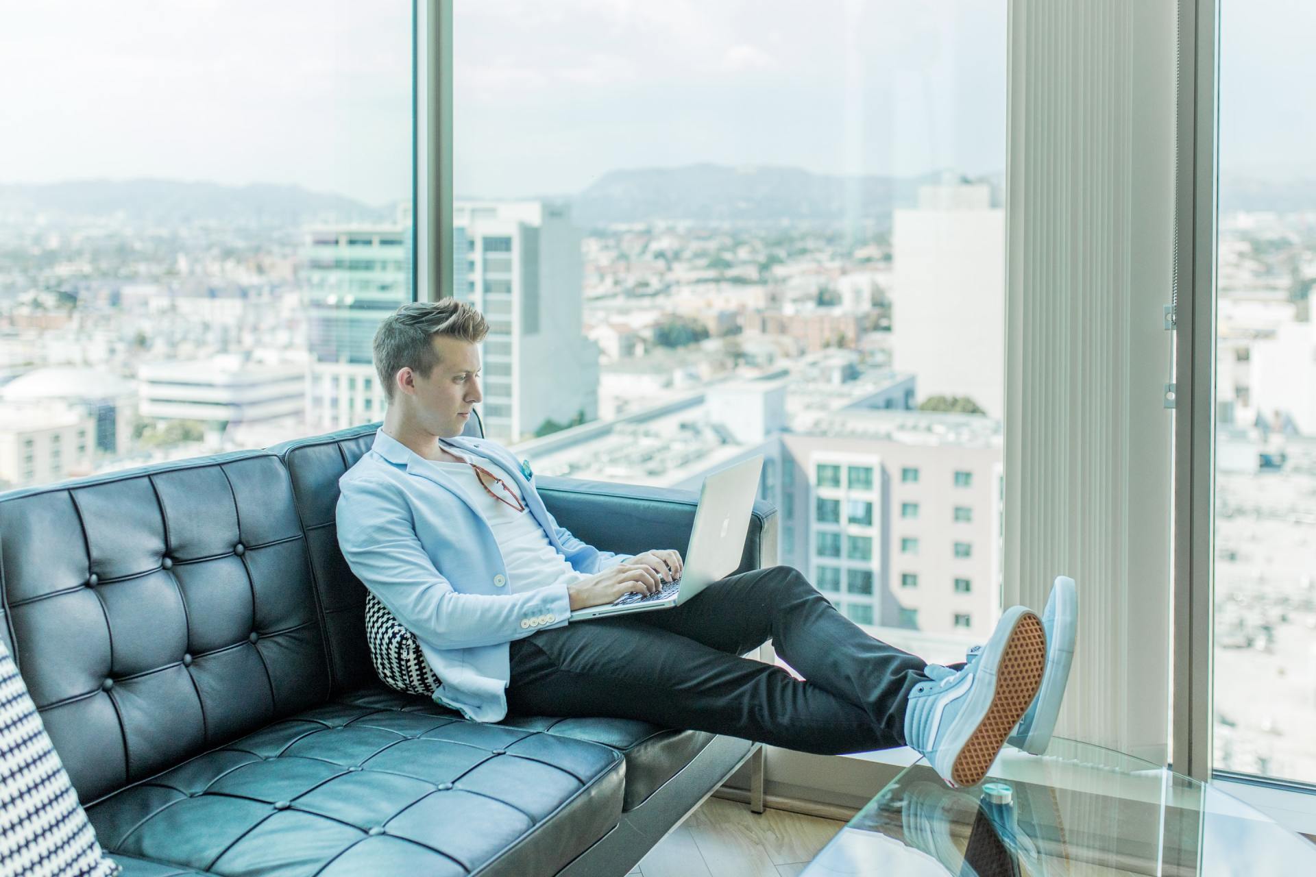 man sat on a sofa in high rise building on his laptop with feet up on glass table
