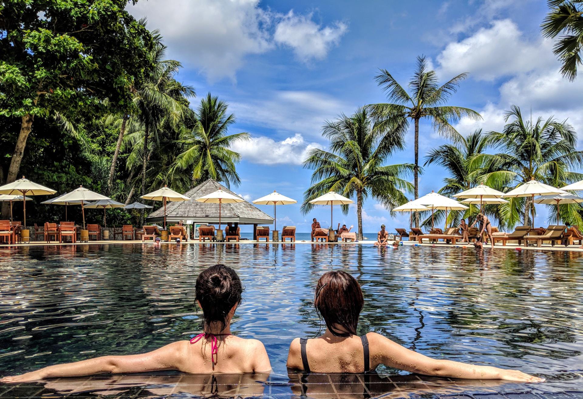 Two people relaxing in a pool with palm trees, umbrellas, and a bright beach resort beyond.