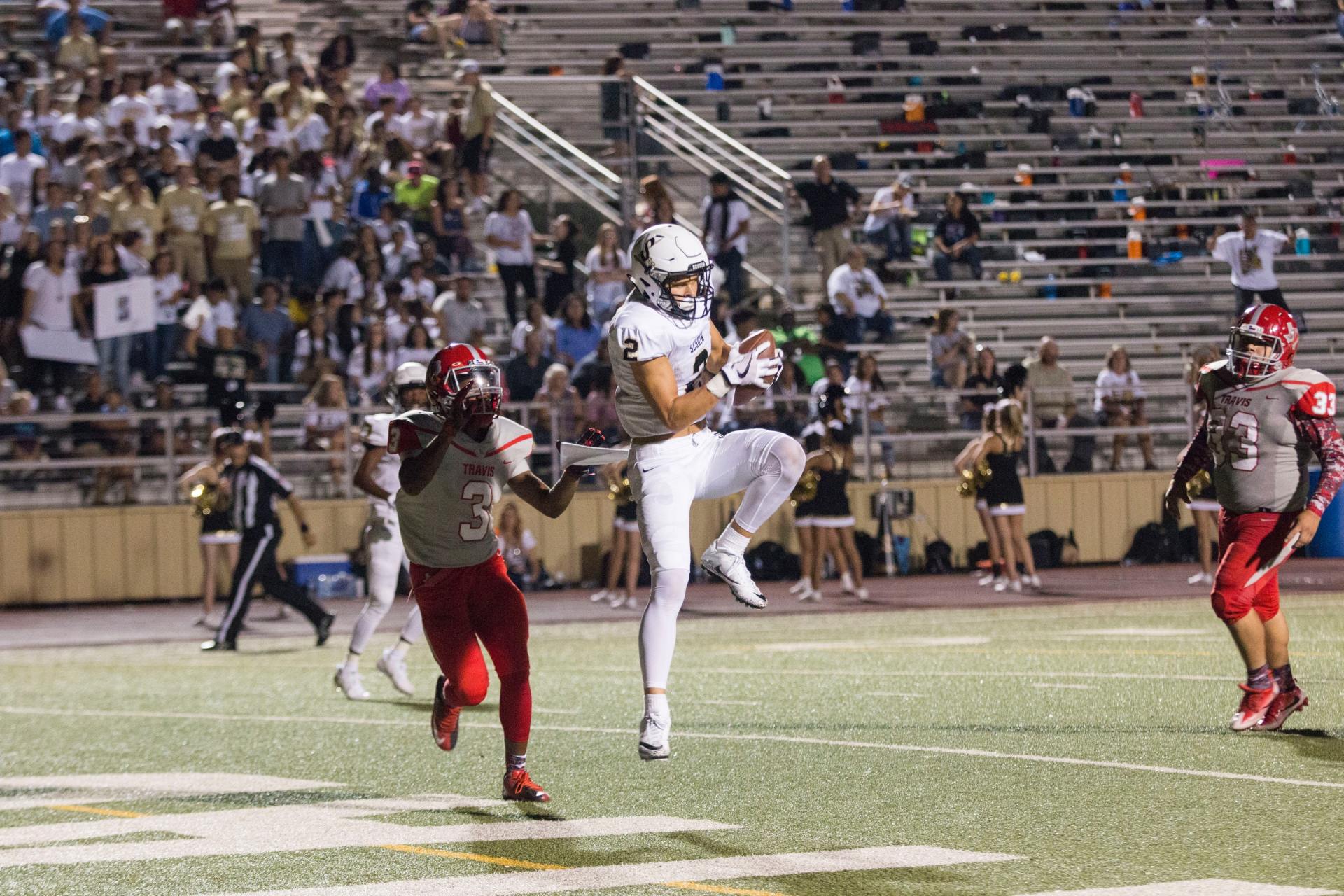 Football player in white jumps to catch ball, another player in red nearby. Stadium crowd in background.