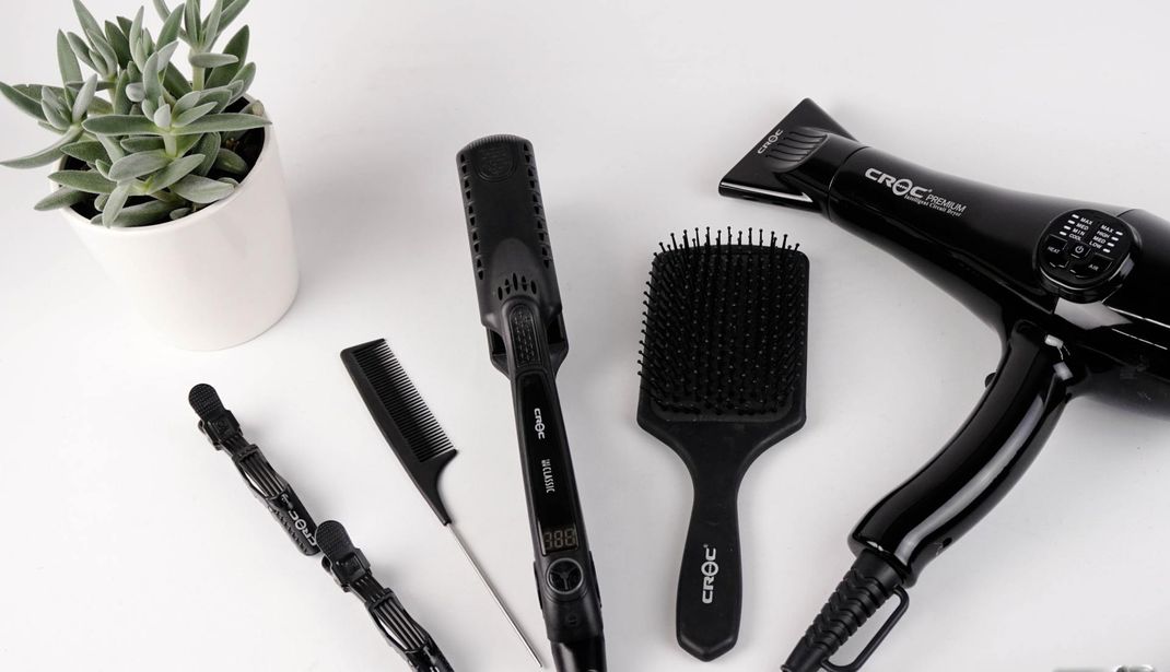 Black hair styling tools including a blow dryer, brushes, a flat iron, and hair clips beside a succulent on a white surface.