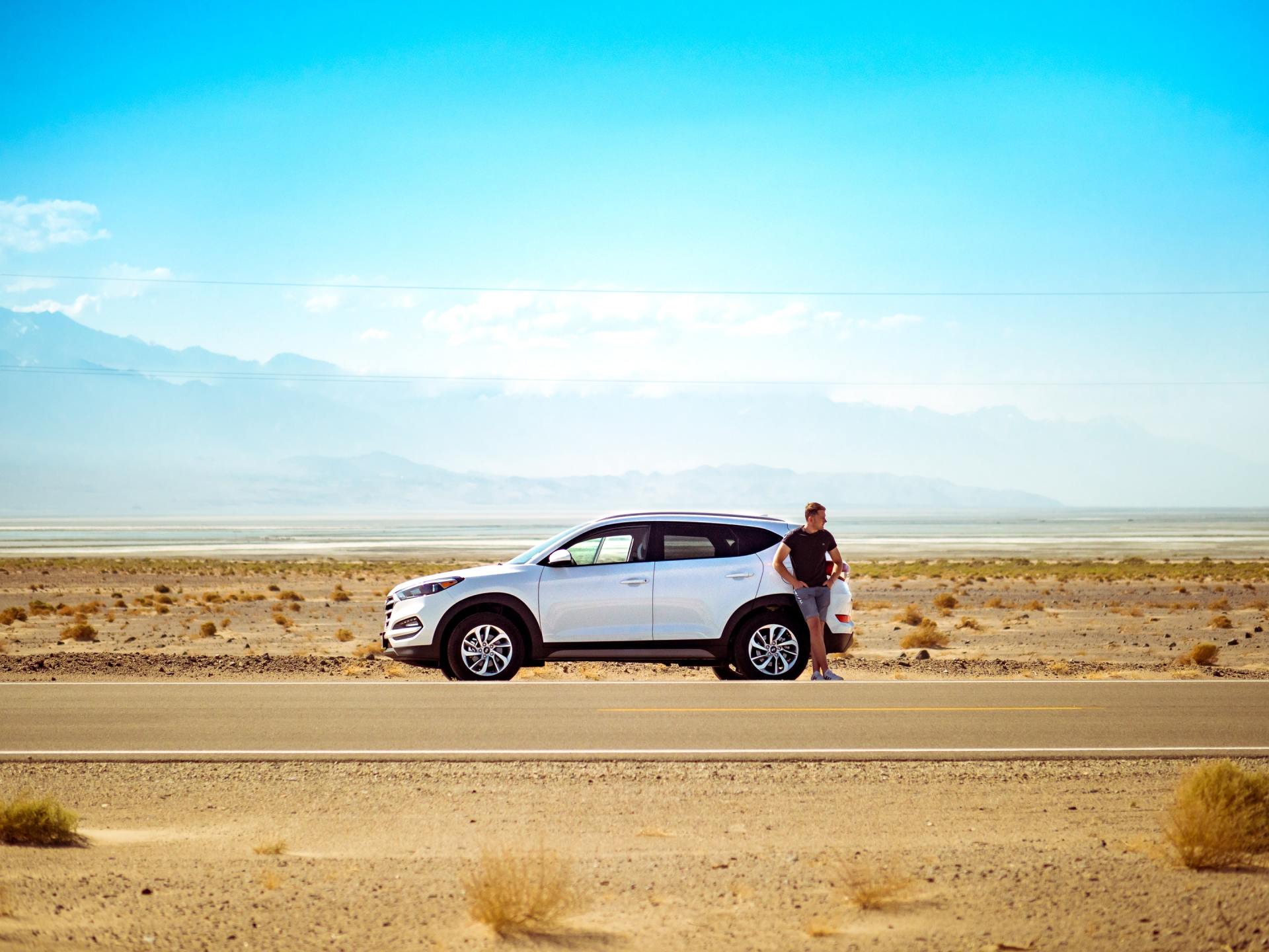White SUV parked on a desert road with a person leaning against it; mountains in the distance under a blue sky.