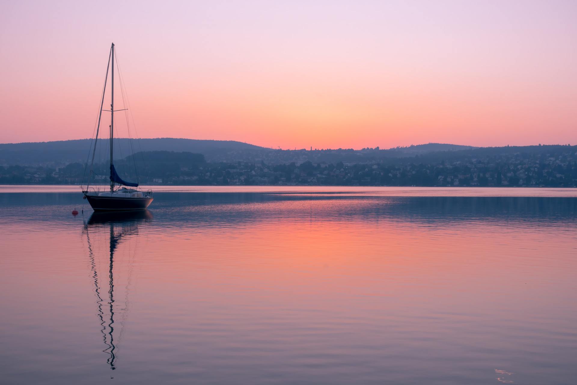 Sailboat on calm pink water at sunset, with a distant shoreline and soft sky reflection