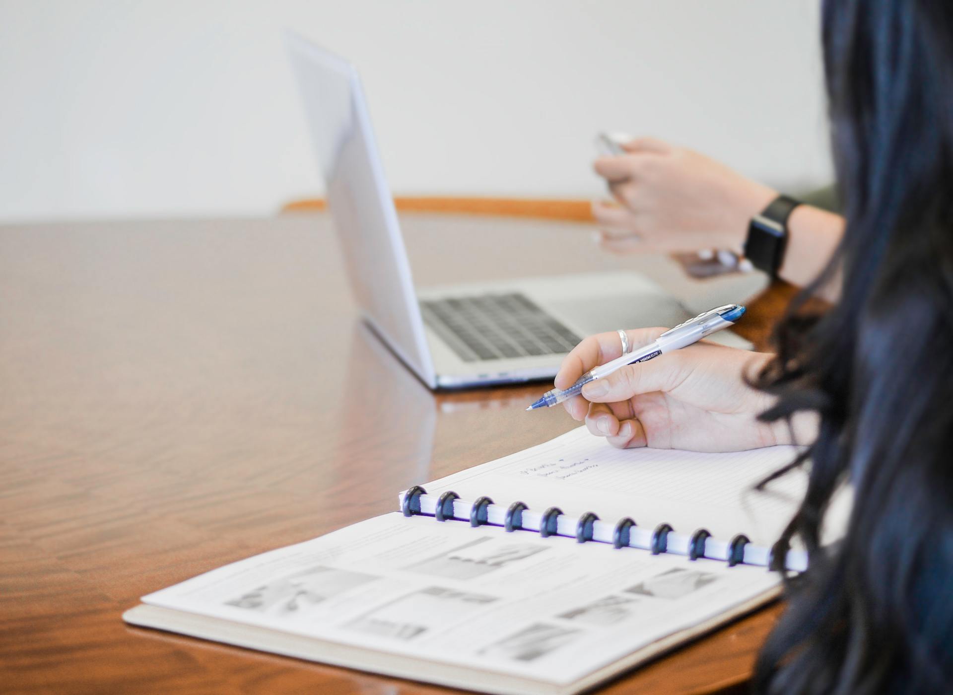 A woman is writing in a notebook while using a laptop computer.