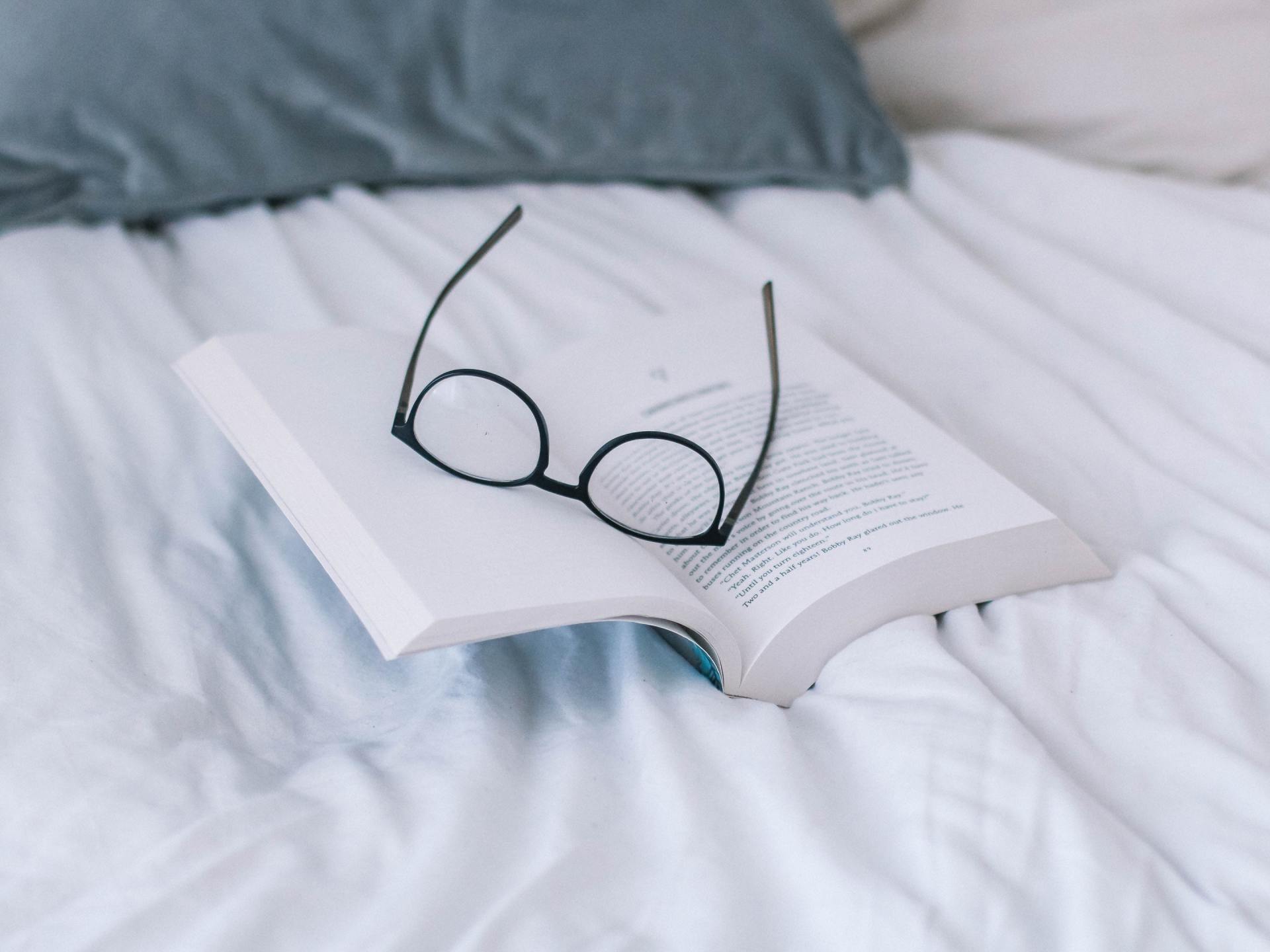 A pair of glasses rests on top of an open book on a bed