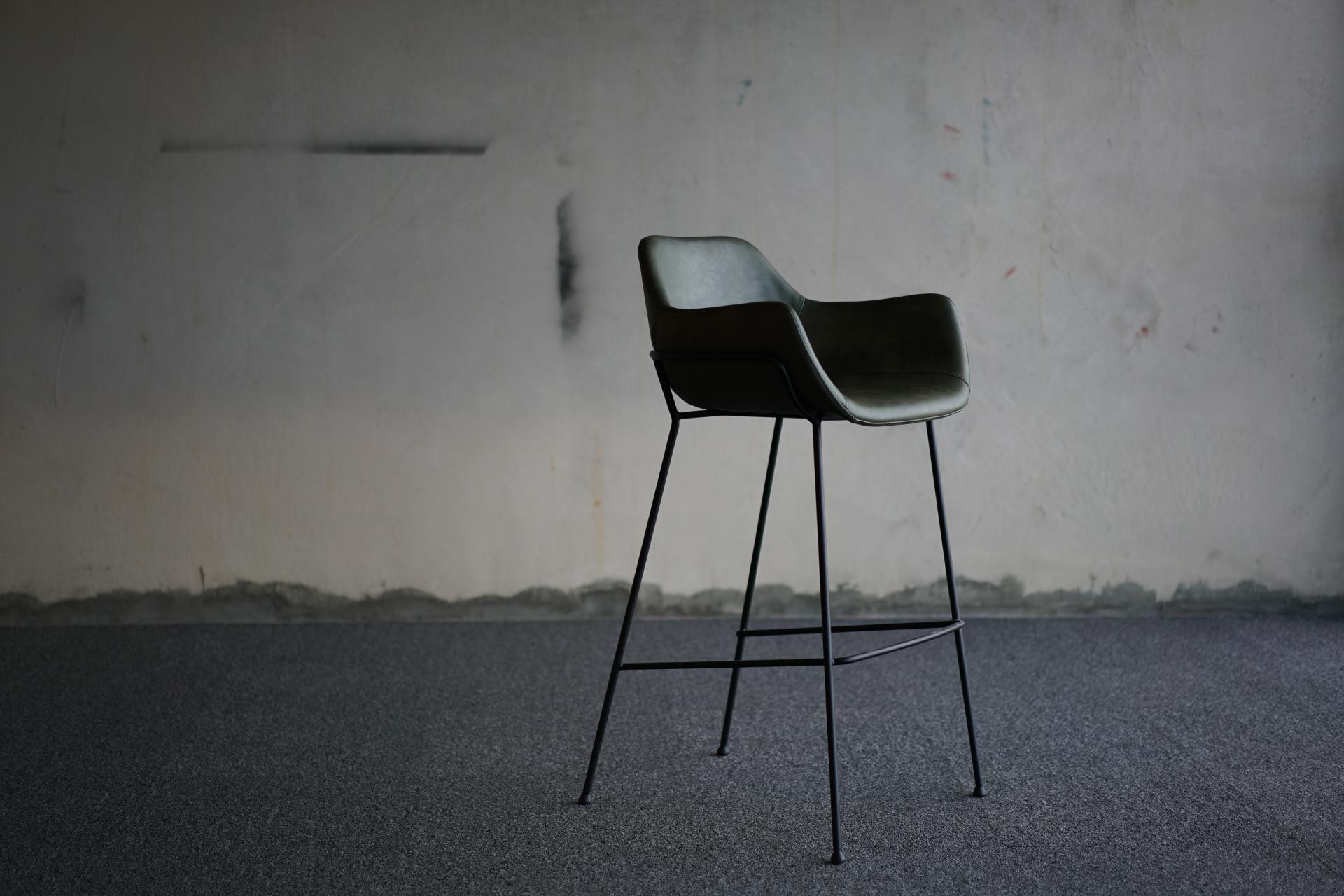 Uno sgabello da bar moderno, di colore scuro e con gambe sottili in metallo, è appoggiato a una parete liscia e usurata, su un pavimento ruvido.