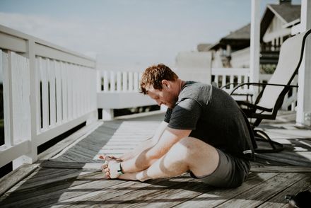 Person sitting on a wooden deck, stretching with head bowed near white railing and lounge chairs.