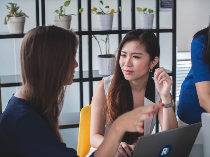 Two women talking at a table with a laptop in a bright office setting