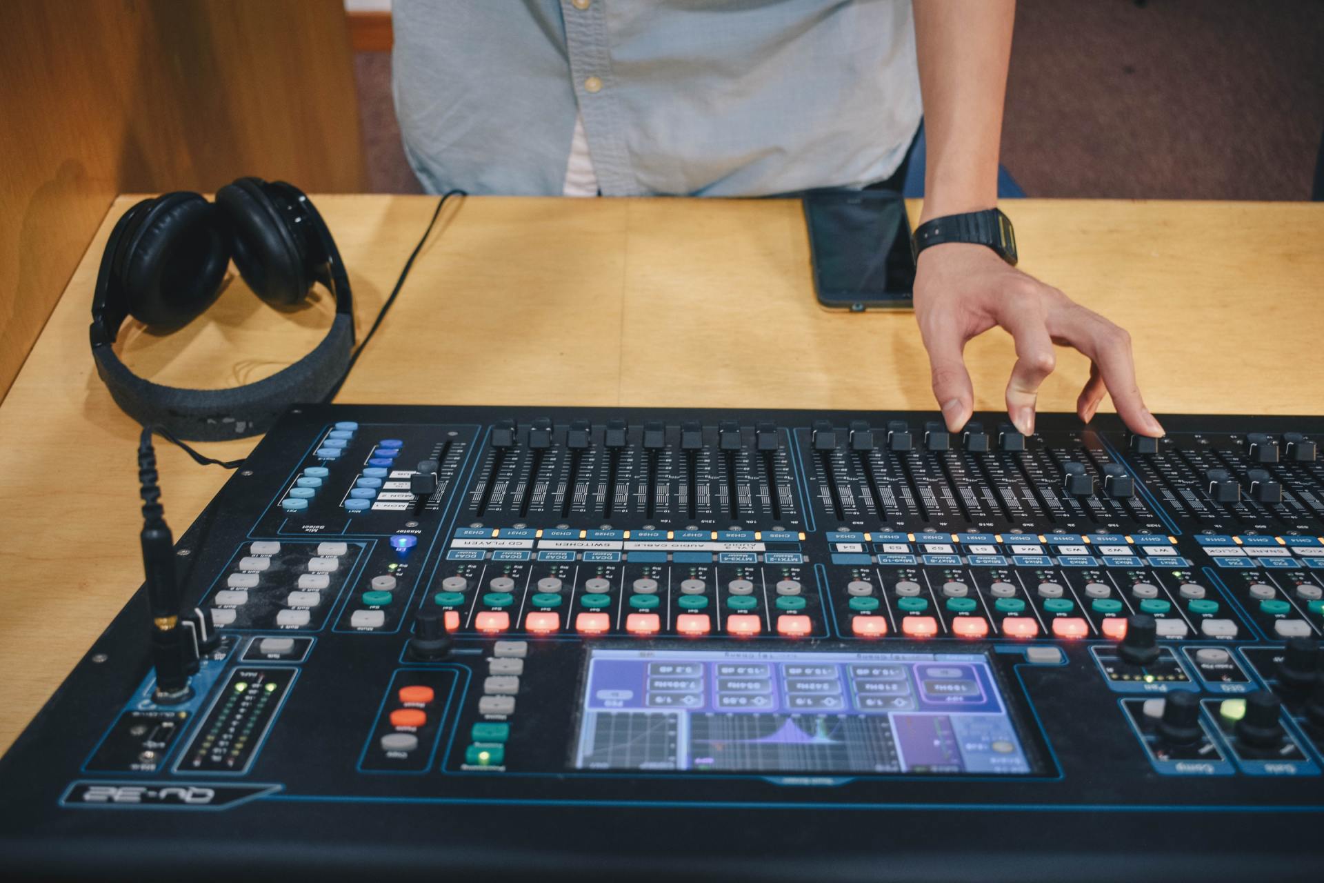Person's hand on a sound mixing console with multiple faders and a screen, headphones to the left.