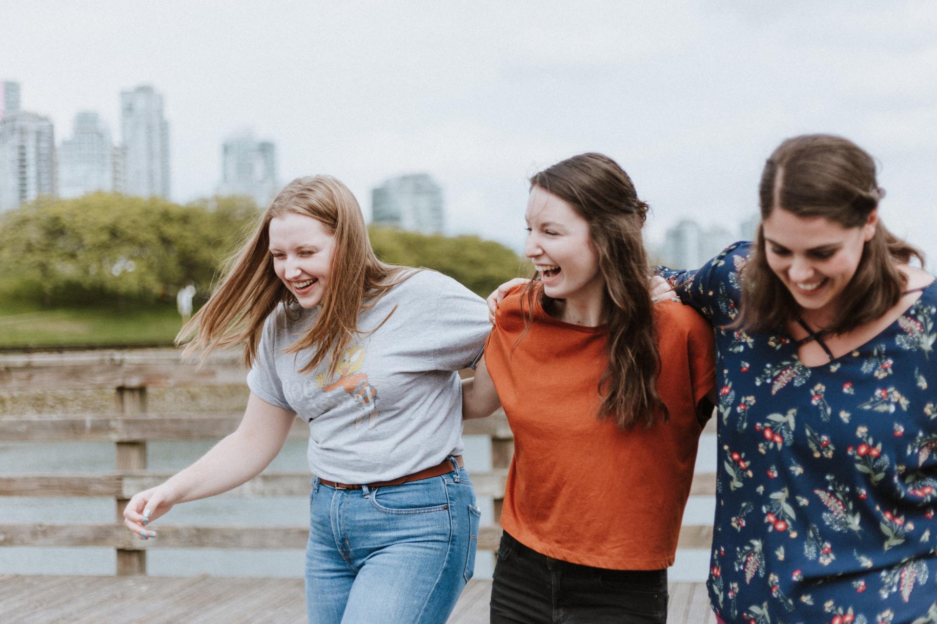 Three friends laughing and walking outdoors by a waterfront railing