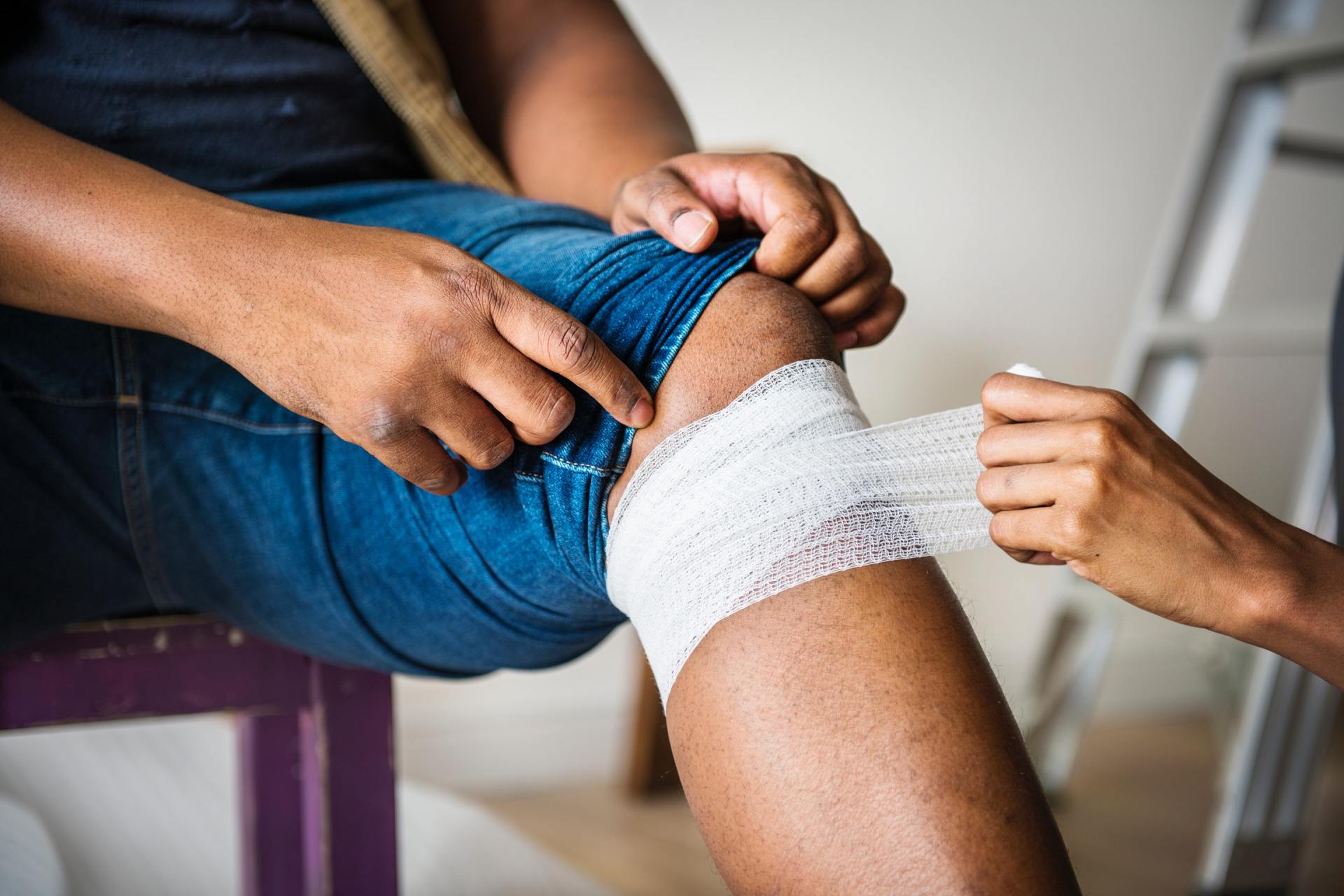 Person's knee being bandaged by another person; indoors, person seated on a purple chair, wearing jeans.