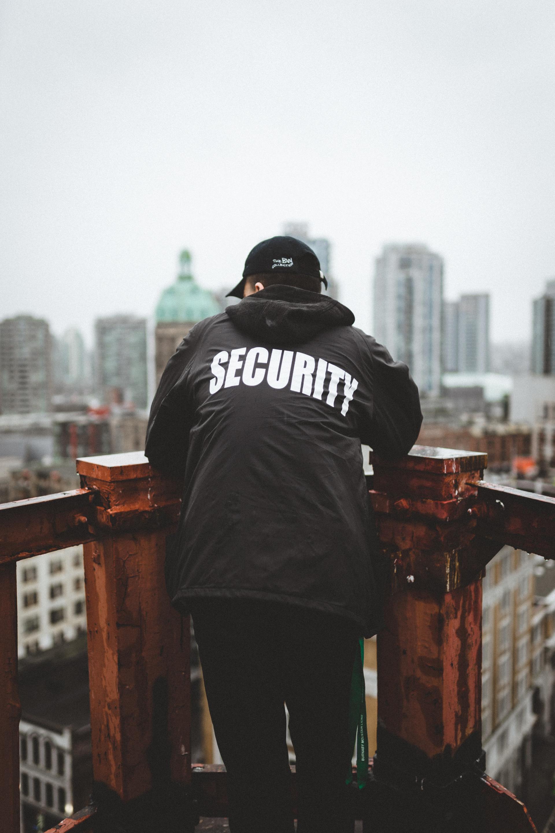 Security guard in a black jacket stands on a rooftop, looking over a city skyline.