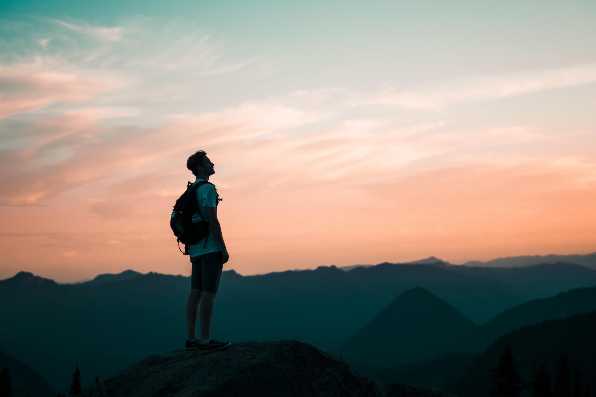 Man with backpack stands atop a mountain, gazing at colorful sunset. Mountains in background.