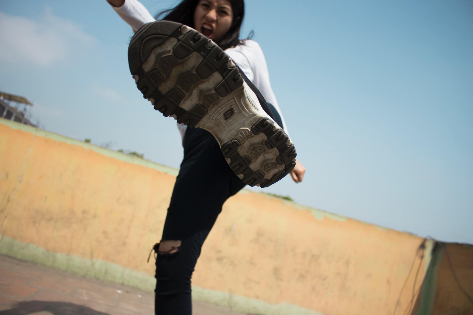 Woman kicking towards the camera, sole of shoe prominent. Blue sky, tan wall, and black pants.