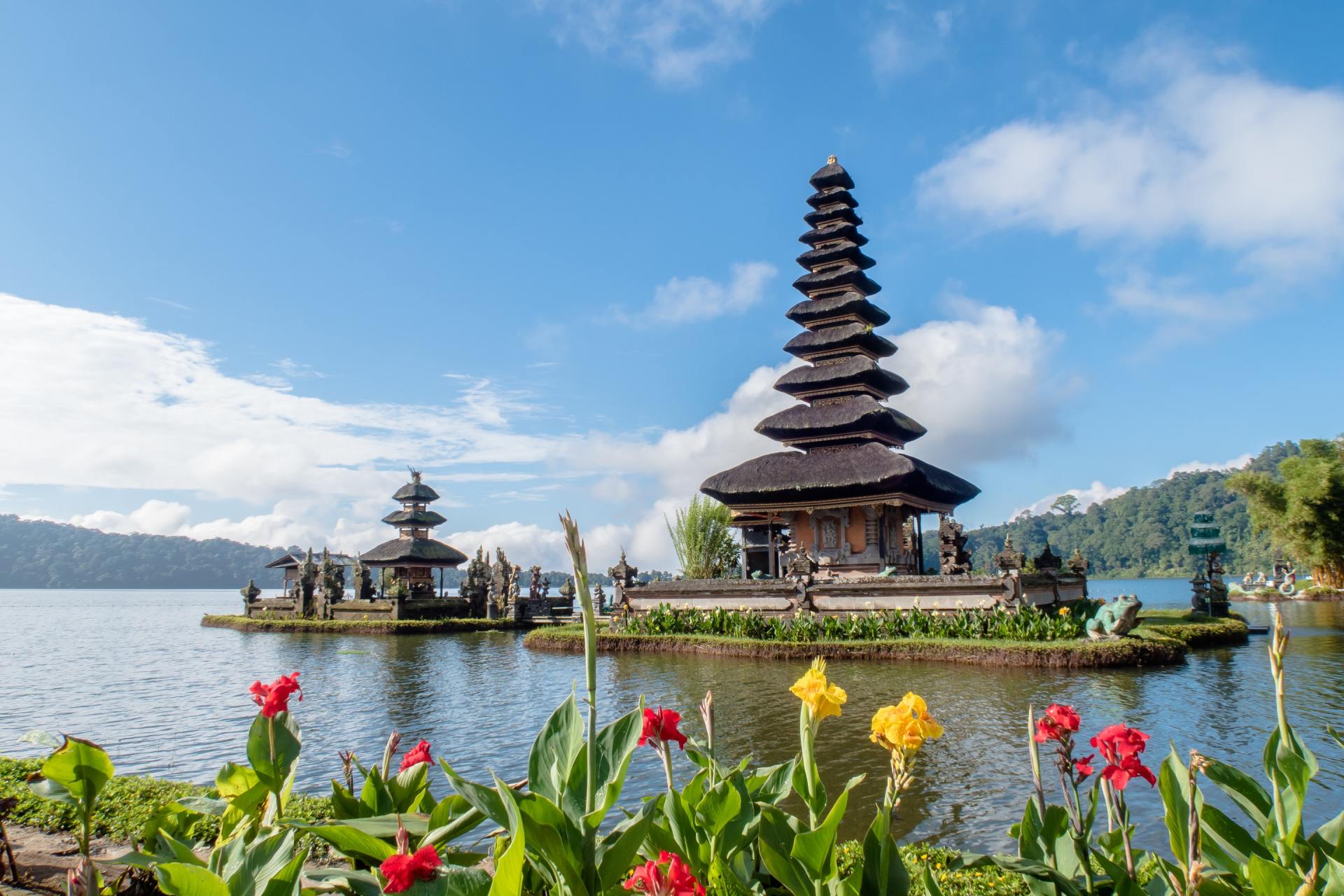 Balinese temple on a lake with tiered roofs, colorful flowers in foreground, blue sky.