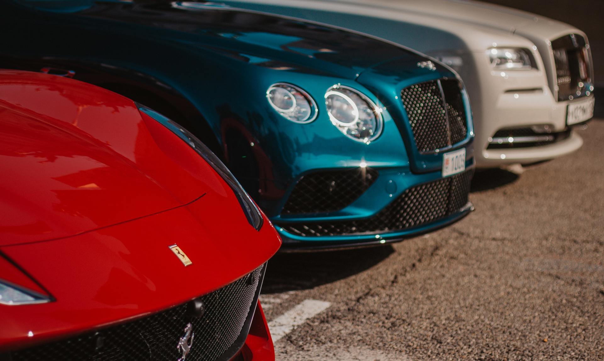 Three luxury cars lined up, including a red sports car and teal and silver Bentleys, on pavement.