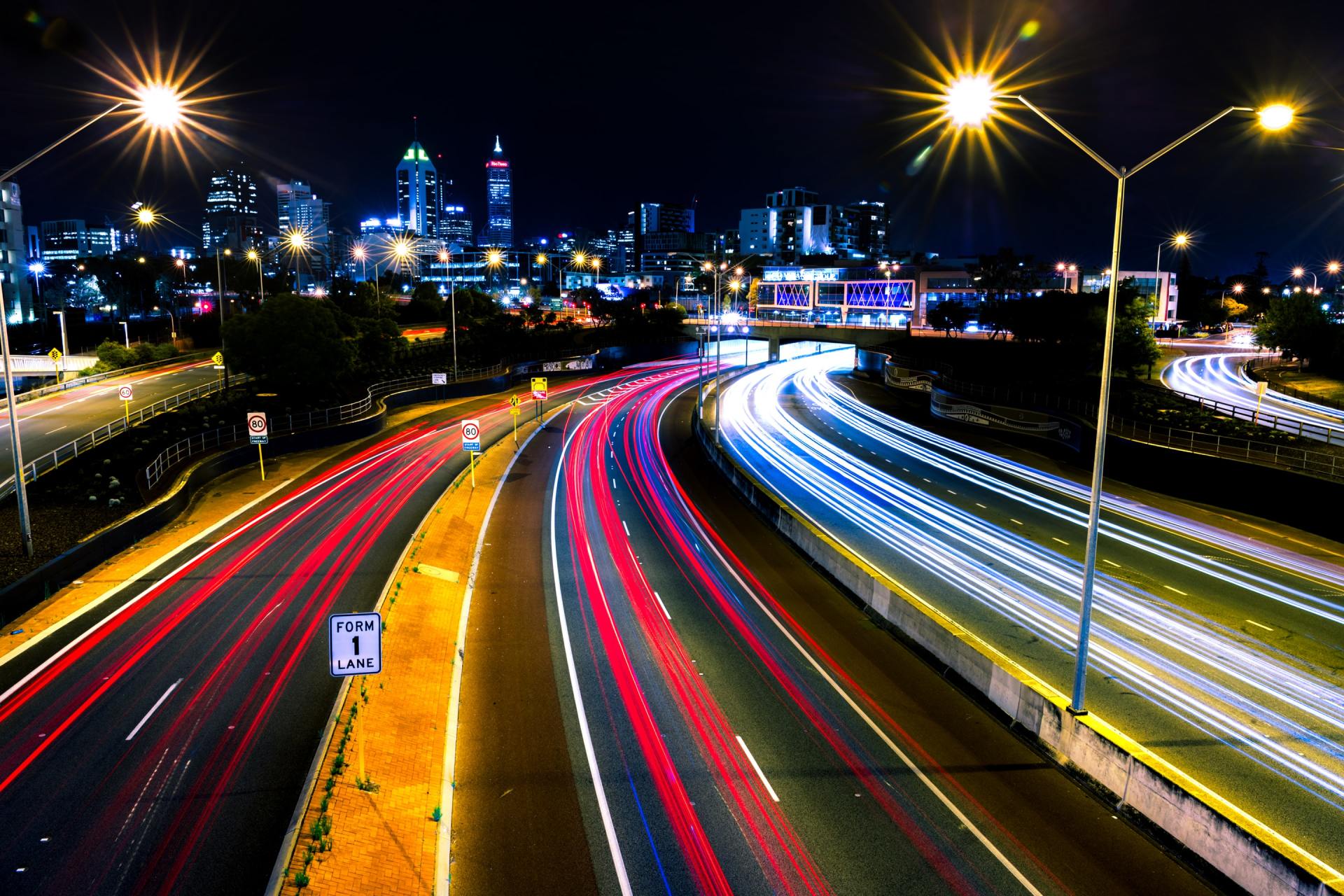 Night highway with blurred red and white car light trails, city lights in background.