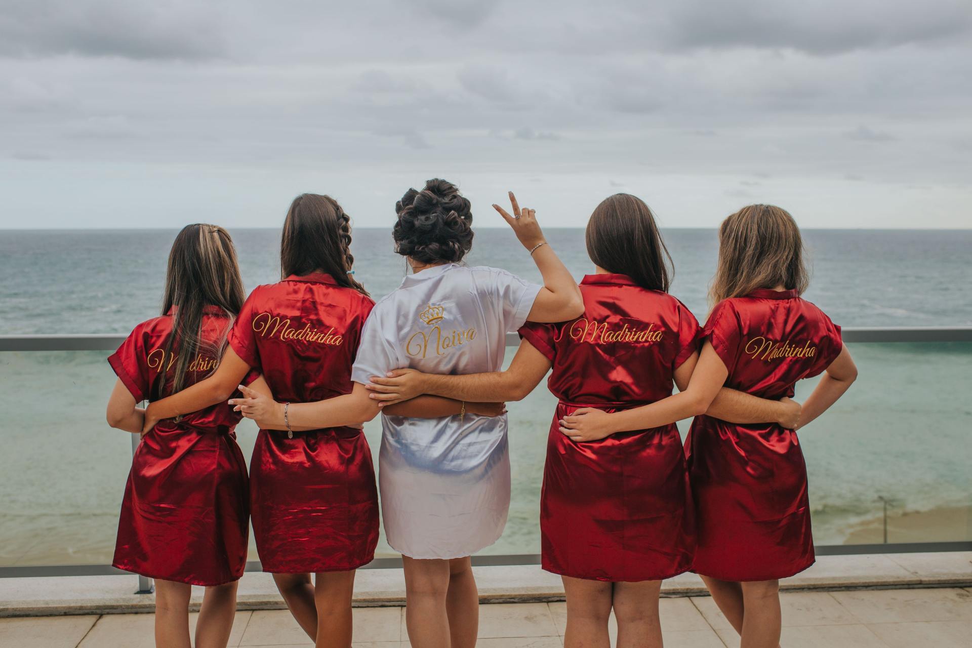 A bride and her bridesmaids are standing on a balcony looking at the ocean.