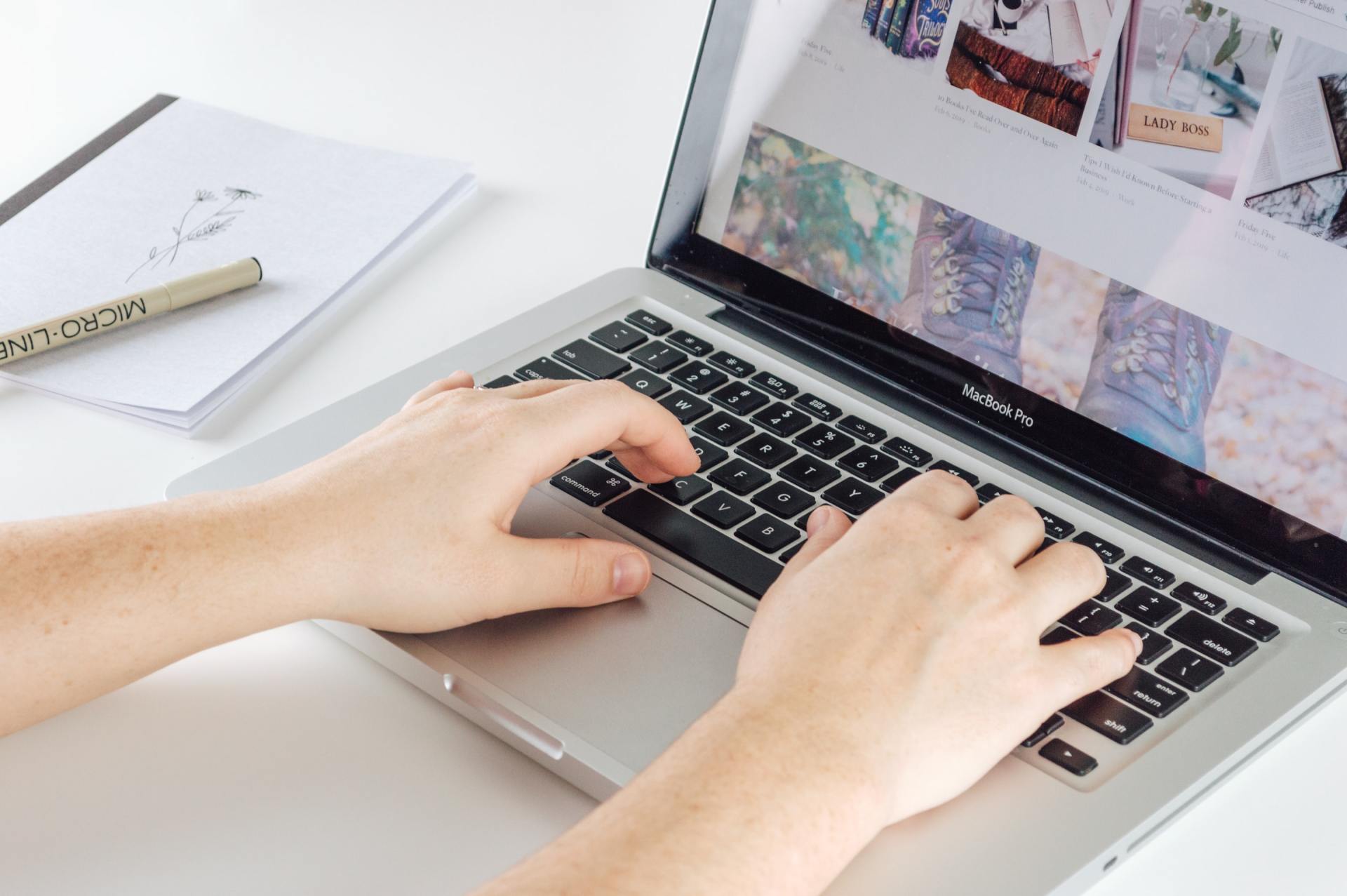 Hands typing on a laptop with a website displayed, notebook and pen nearby on a white desk.