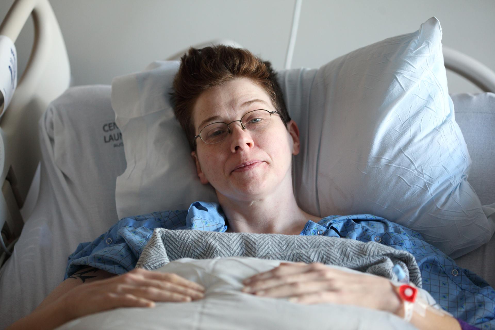Woman in a hospital bed wearing glasses and a hospital gown, with an IV in her arm; she is smiling.