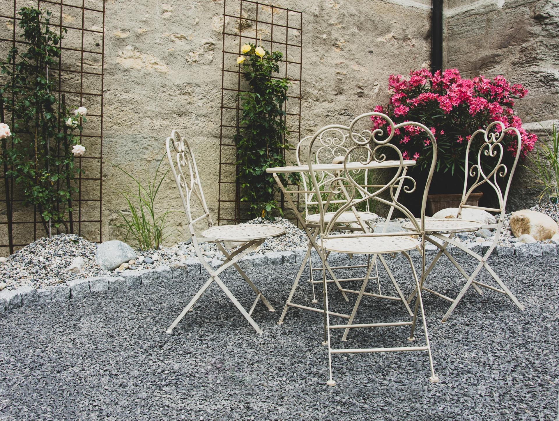 Un piccolo bistrot all'aperto con sedie in ferro battuto color crema su un patio di ghiaia, addossato a un muro di pietra ricoperto di rampicanti.