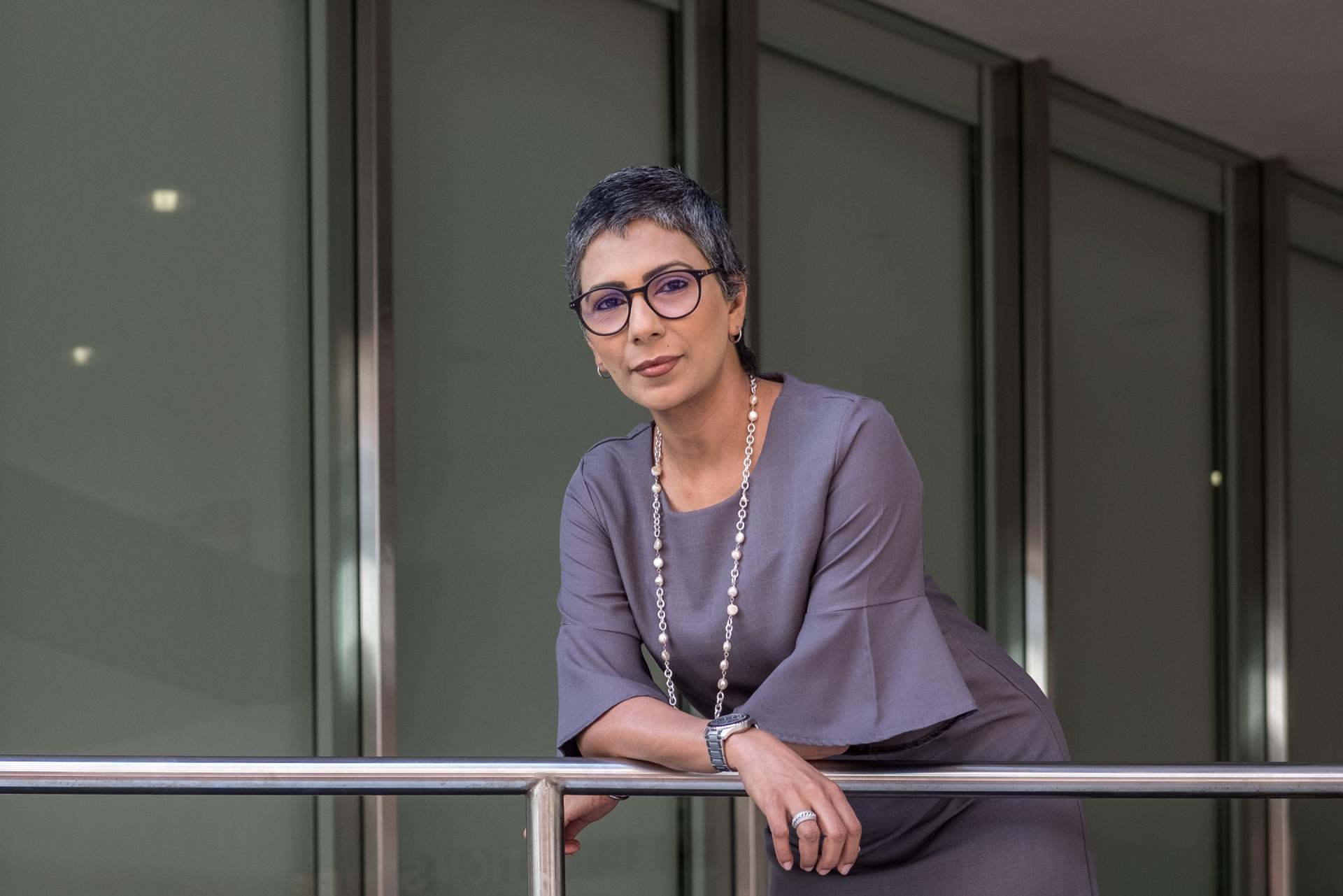 A woman in a gray dress and glasses is leaning on a railing.