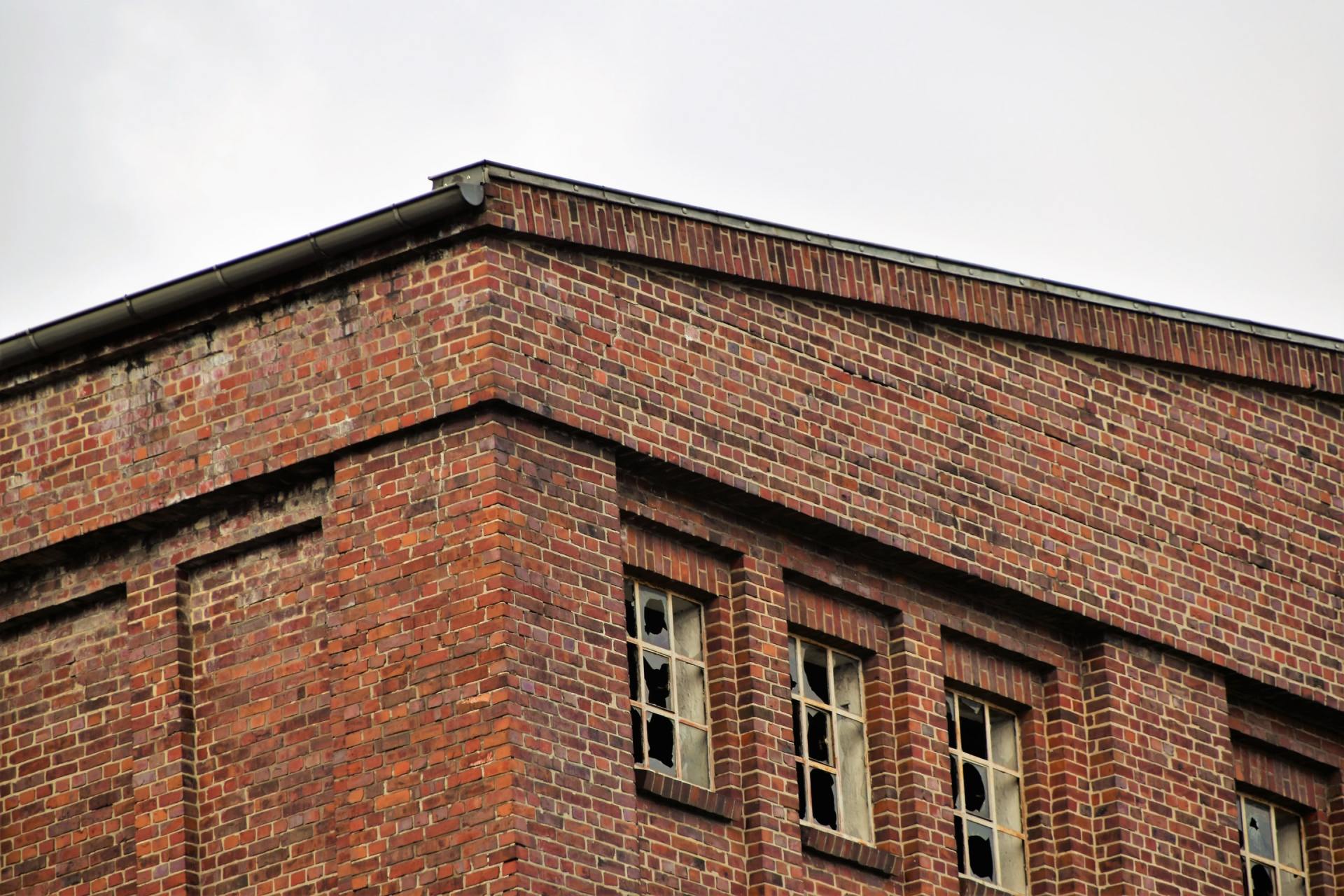The roof of a brick building with broken windows against a cloudy sky.