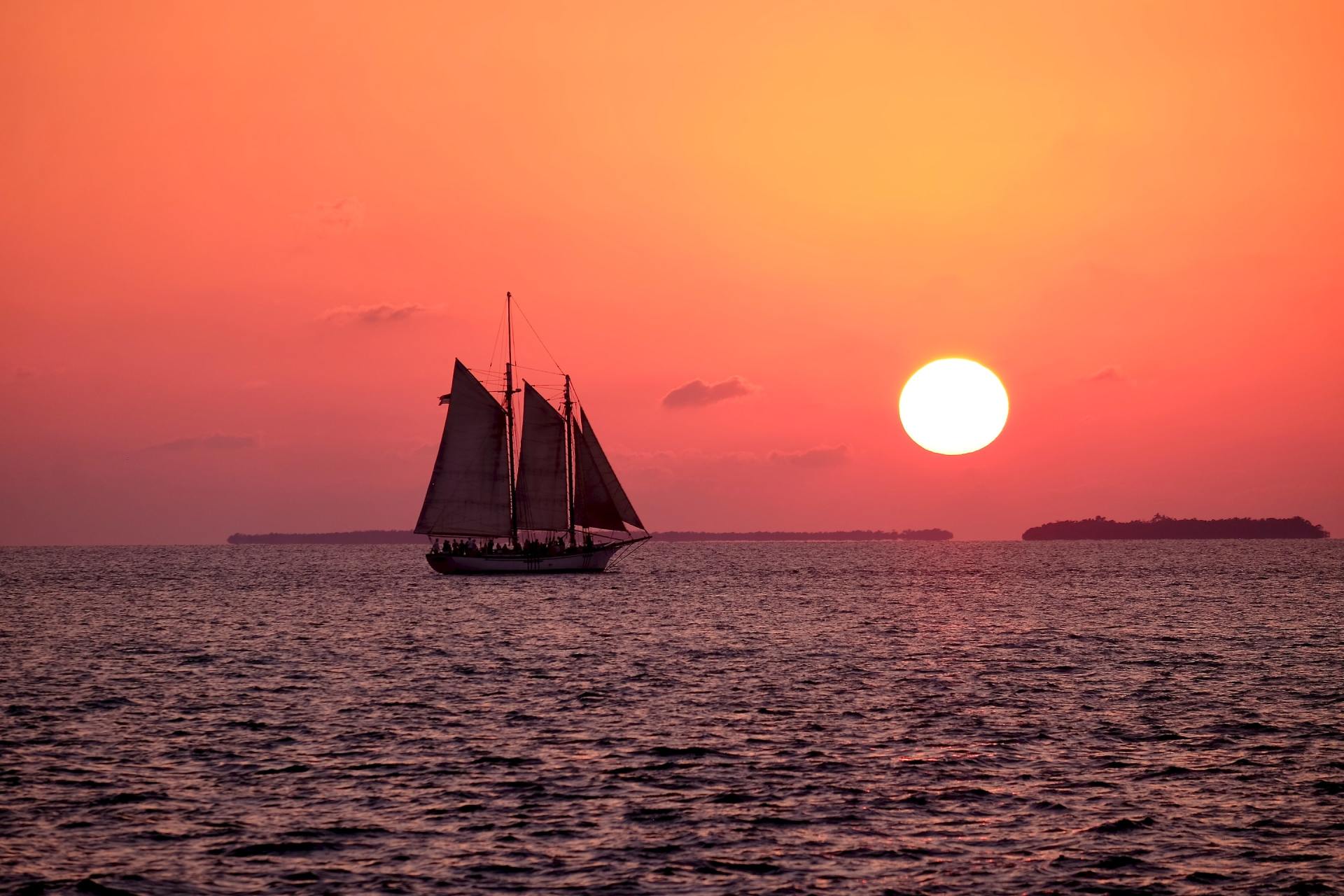 Silhouette eines Segelboots auf ruhiger See bei Sonnenuntergang, mit einer großen orangefarbenen Sonne über dem Horizont