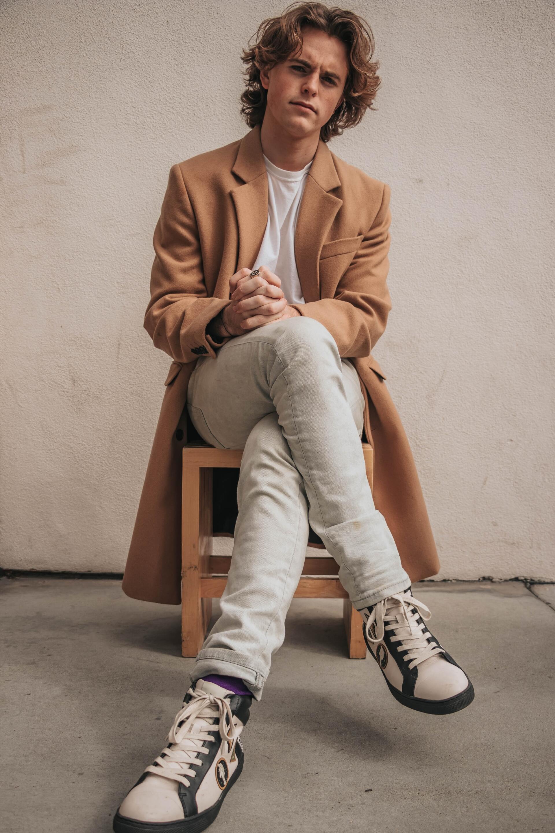 A young man is sitting on a wooden stool with his legs crossed.