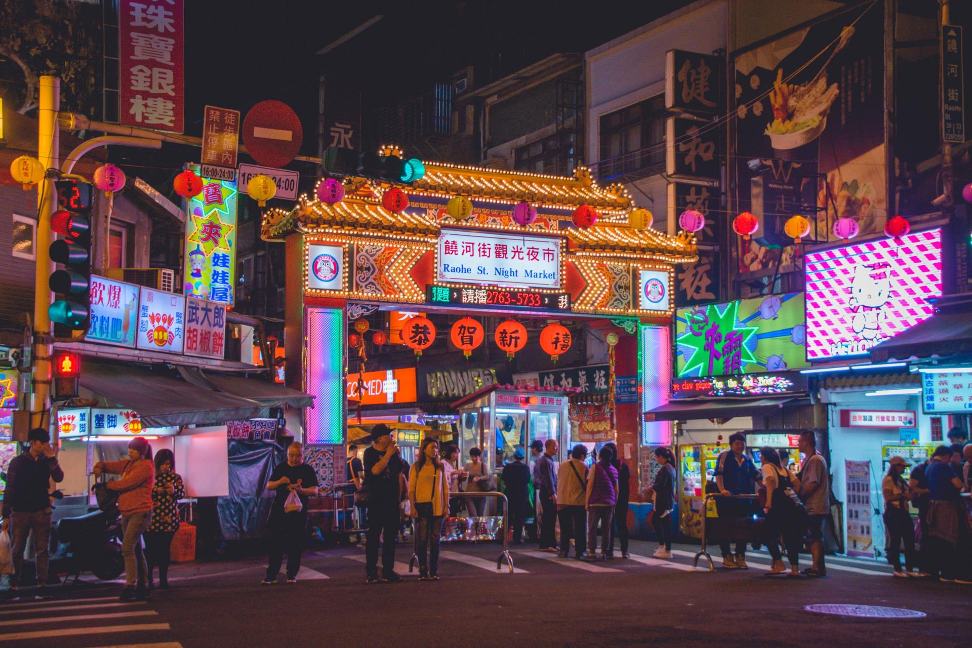 Nighttime street scene in Taiwan, brightly lit with neon signs, food stalls, and people.