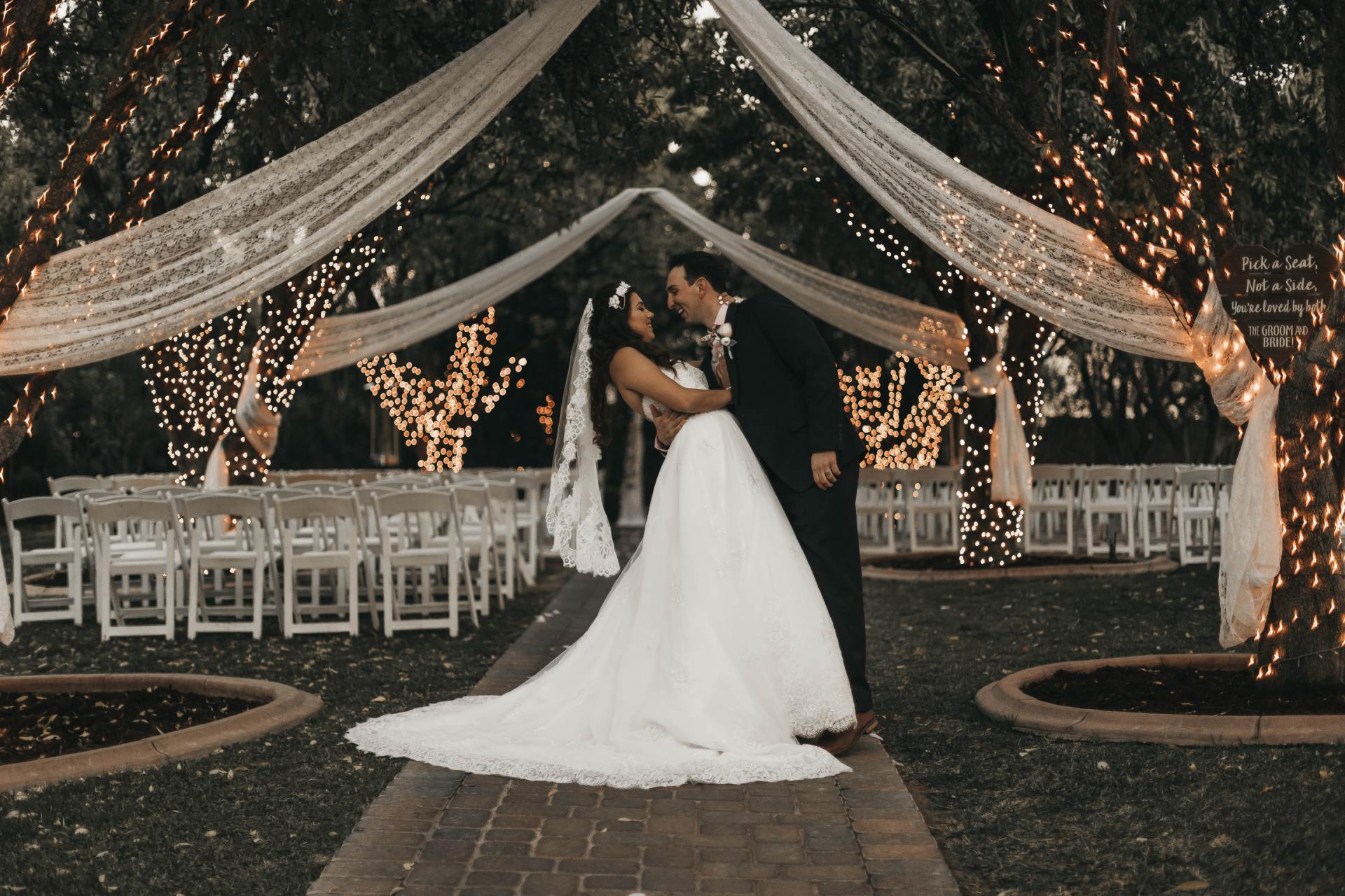 Couple kissing at a wedding ceremony. Bride in white gown, groom in a suit, outdoor setting with draped fabric and lights.