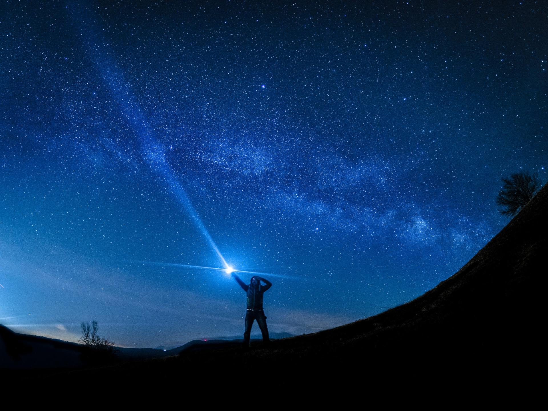 La silueta de una persona de pie en una colina por la noche, apuntando un brillante haz de luz hacia un cielo estrellado.