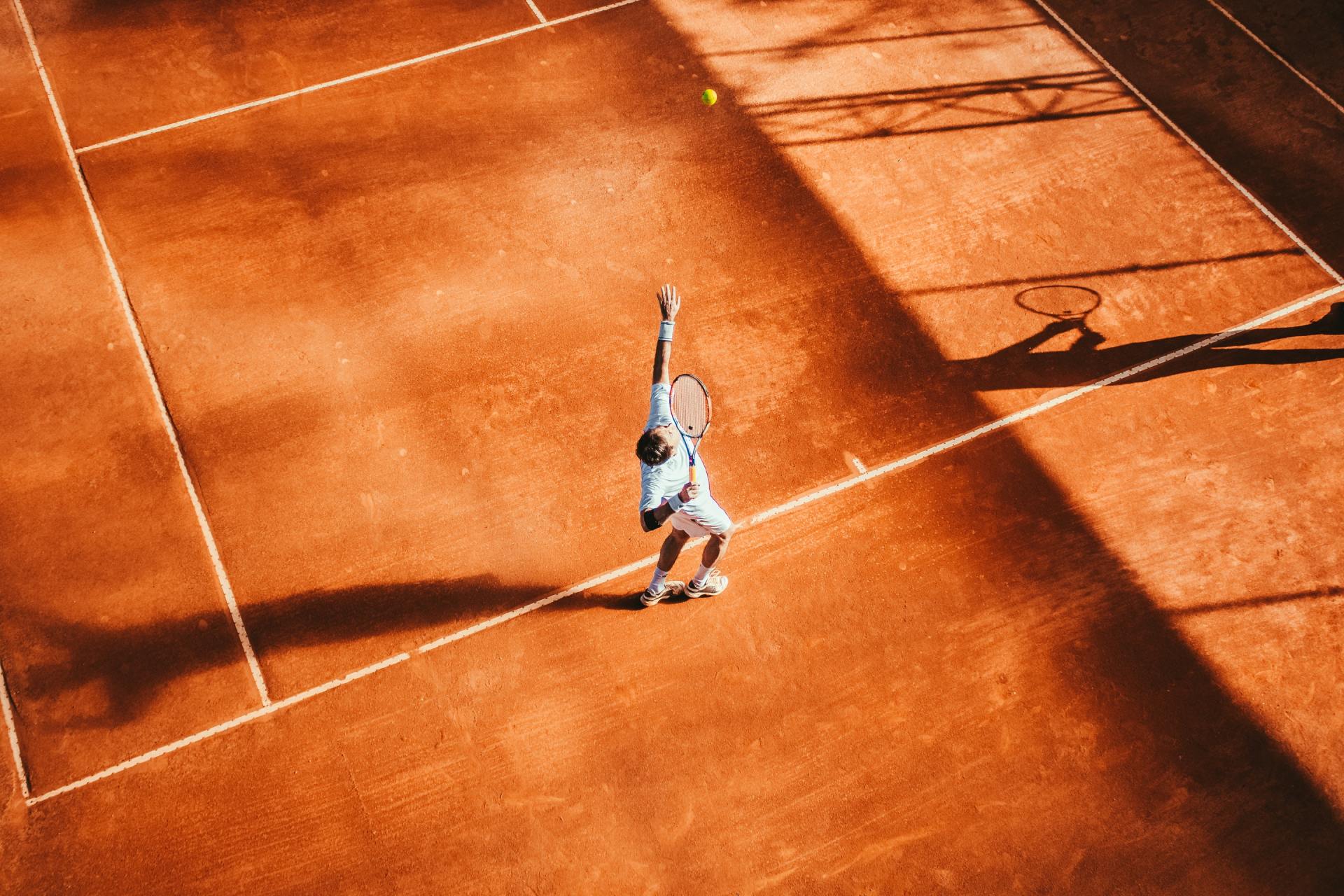 Tennis player serving on a clay court; racquet shadow visible.