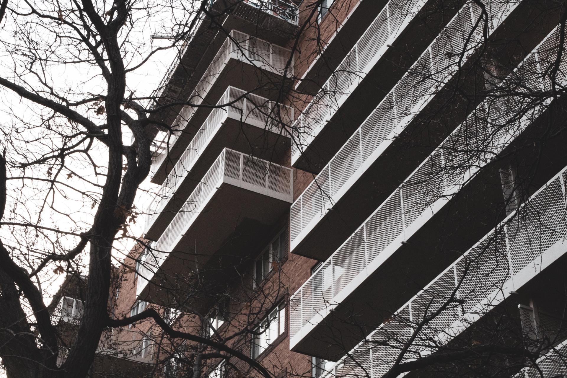 Tall apartment building with stacked balconies and bare tree branches against a cloudy sky